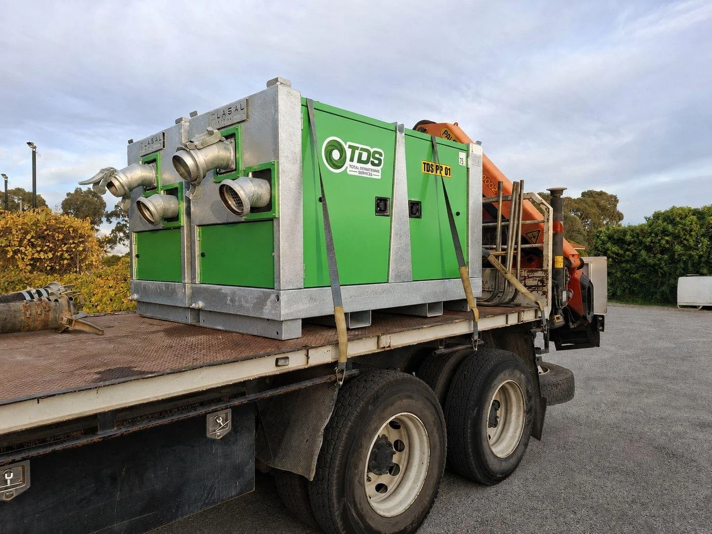 A green industrial pump or compressor mounted on a flatbed truck with metal pipes and hoses, outdoors on a gravel surface with trees and a cloudy sky in the background.