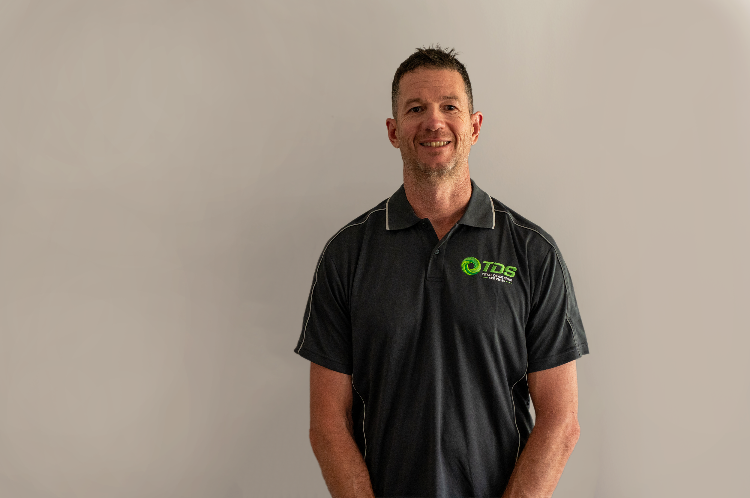 A man wearing a black polo shirt with a green logo standing against a plain light-colored wall.