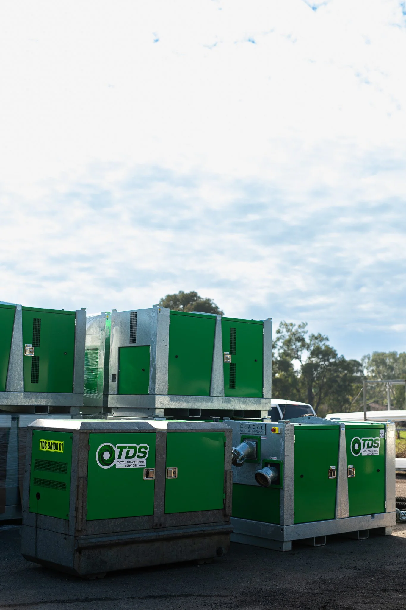 Green and gray industrial machines labeled TDS, stacked outdoors on pallets, with trees and a partly cloudy sky in the background.