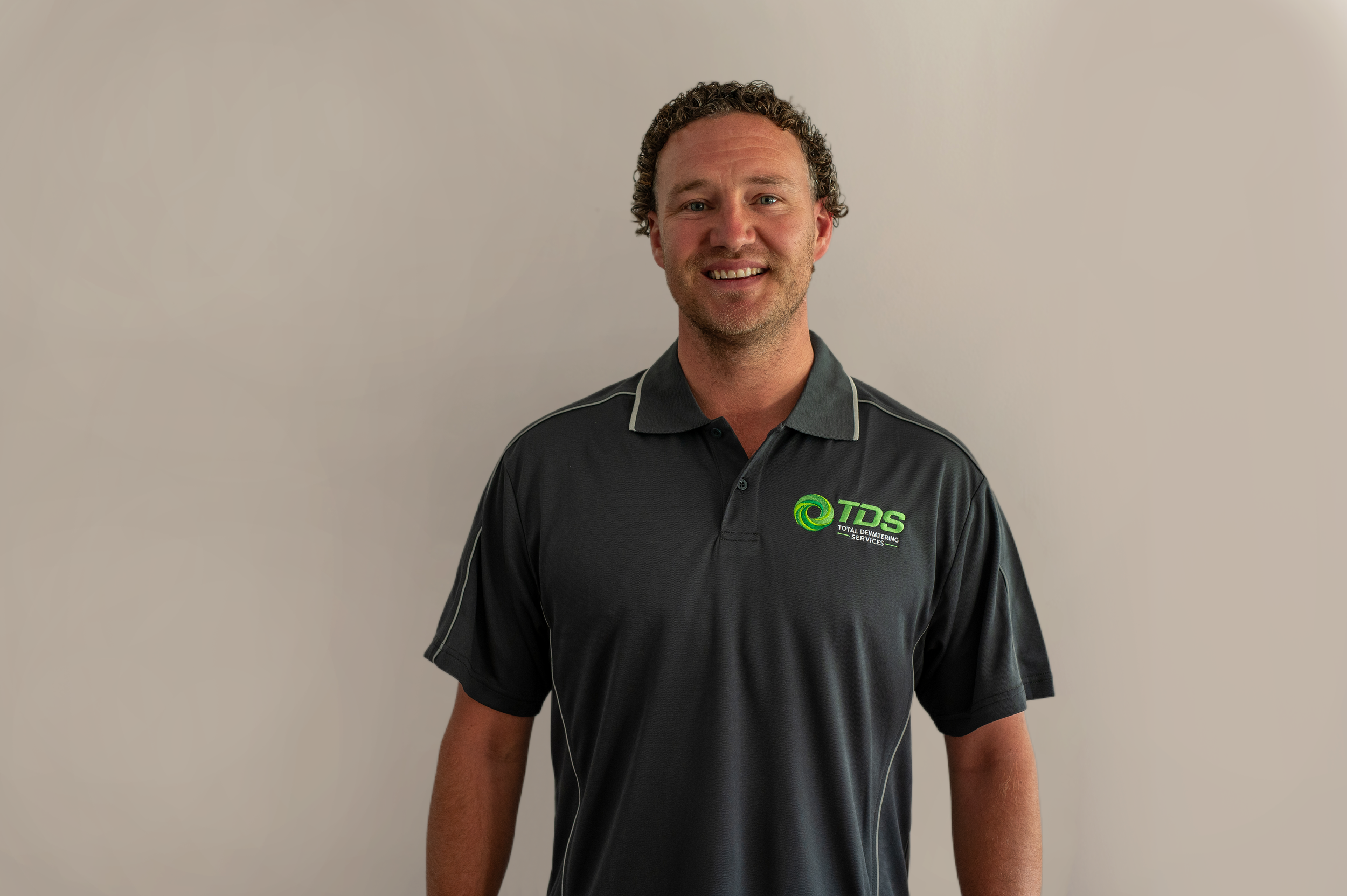 A man standing in front of a beige wall, wearing a black polo shirt with the logo of TDS Total De-Watering Services, smiling at the camera.