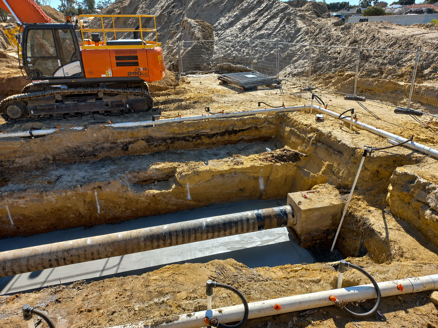 Construction site with an excavator and piping system, excavation trenches, and a chain-link fence in the background.
