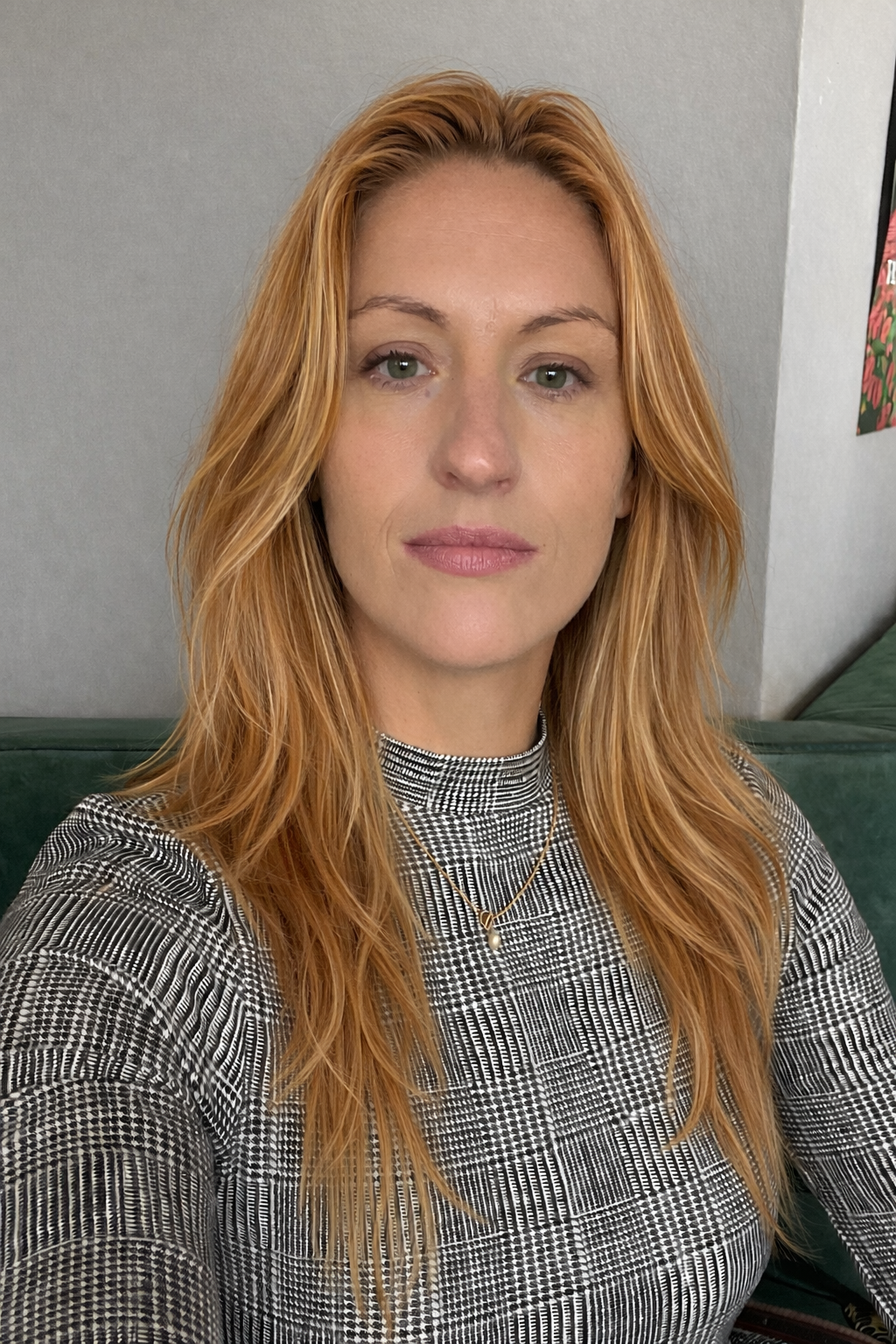 A woman with long, wavy red hair, wearing a houndstooth patterned top, a pearl necklace, and standing in front of a plain wall with a piece of colorful art partially visible.