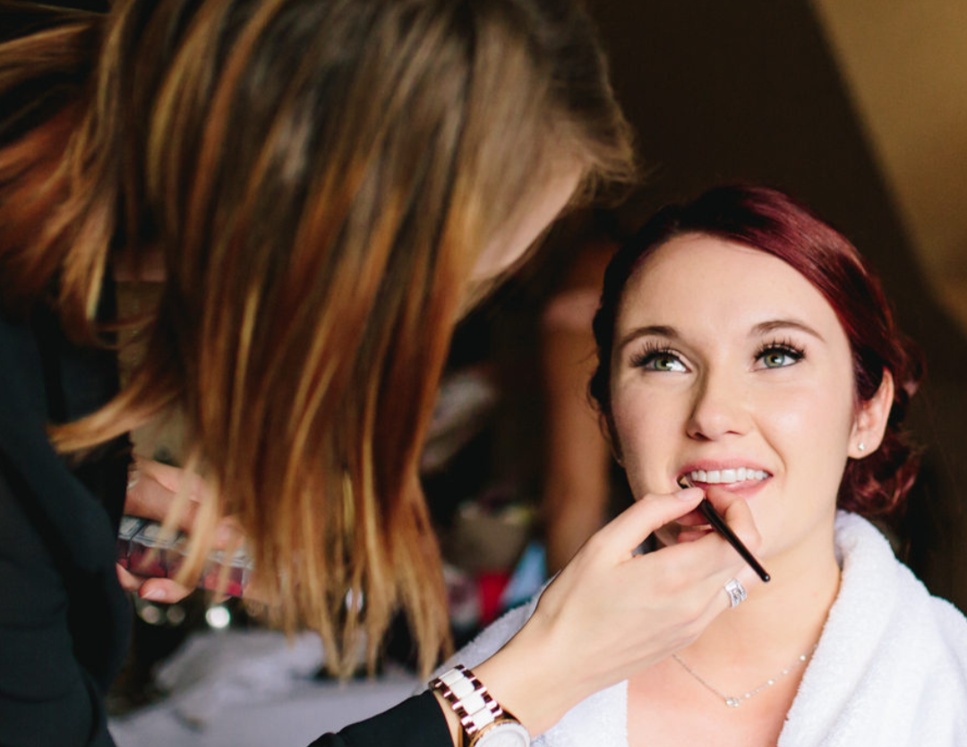 Makeup artist applying lipstick to a woman with red hair in a well-lit room.