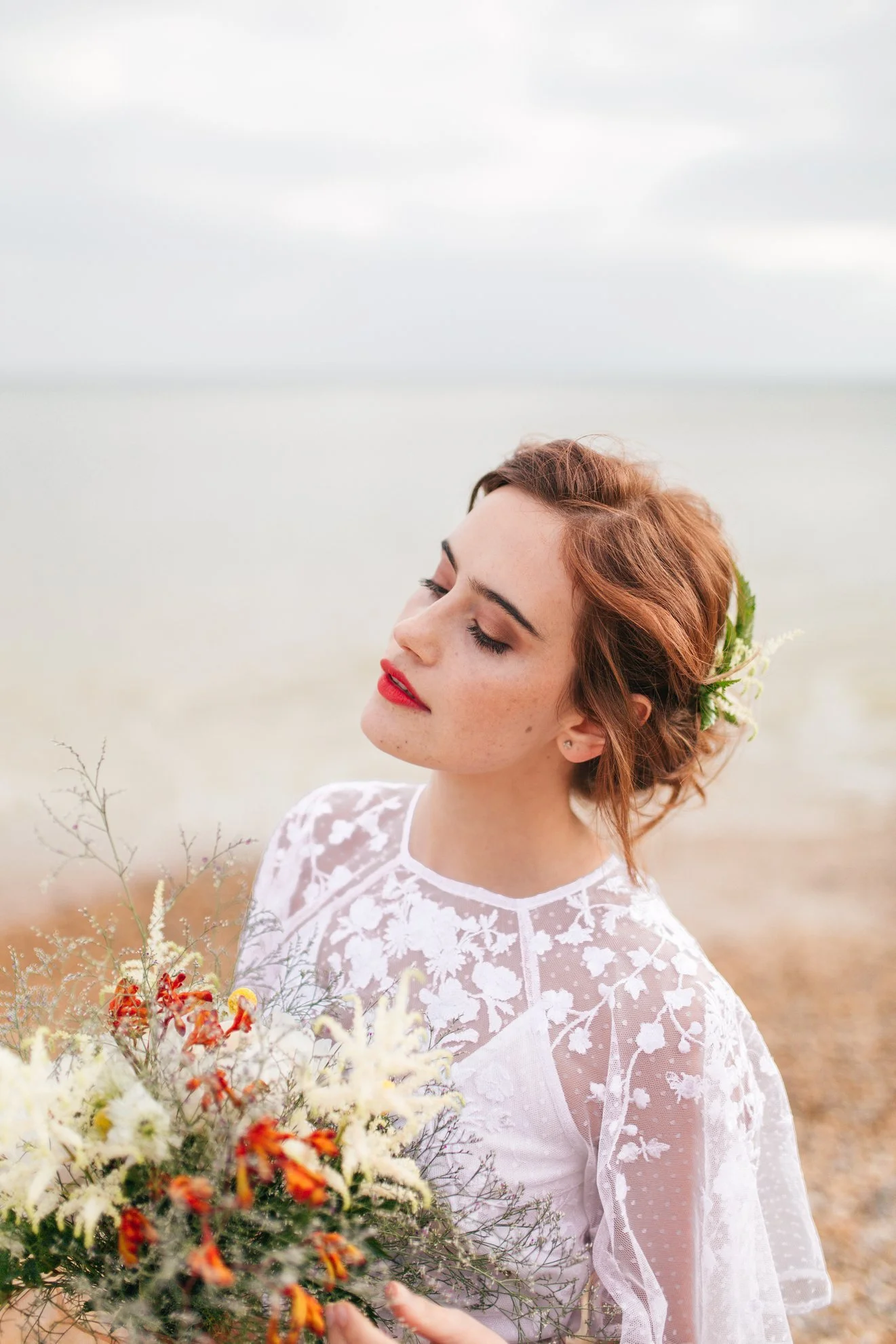 A woman with red hair and closed eyes holding a bouquet of flowers outdoors near a body of water.