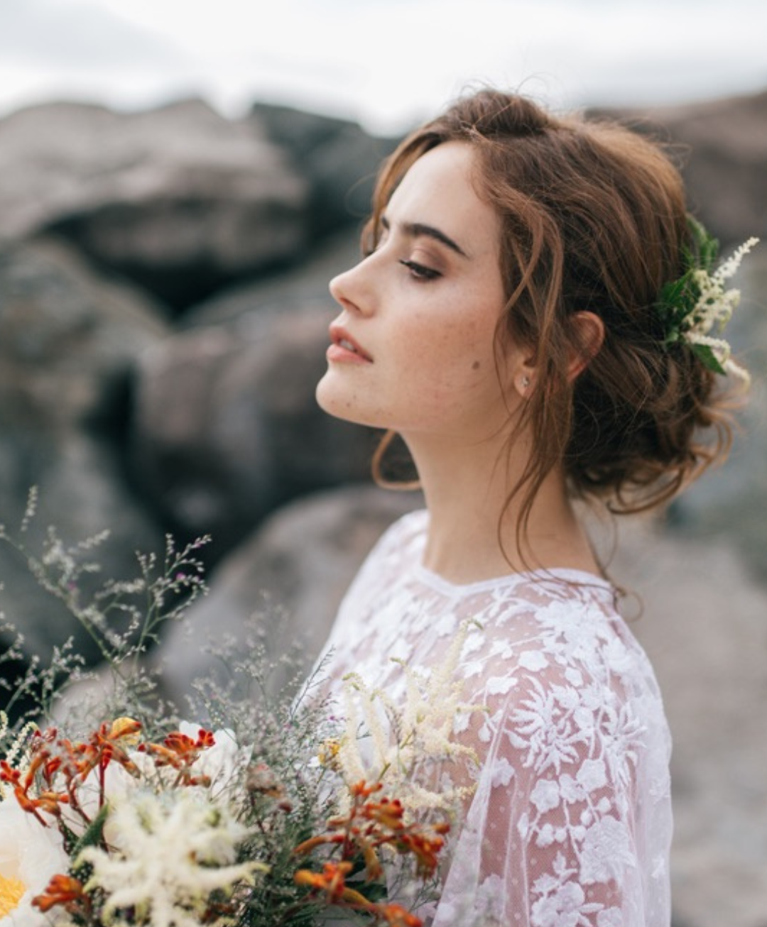 A woman with auburn hair in a loose, flowy hairstyle with flowers, wearing a white lace dress, standing outdoors near rocks, holding a bouquet of wildflowers.