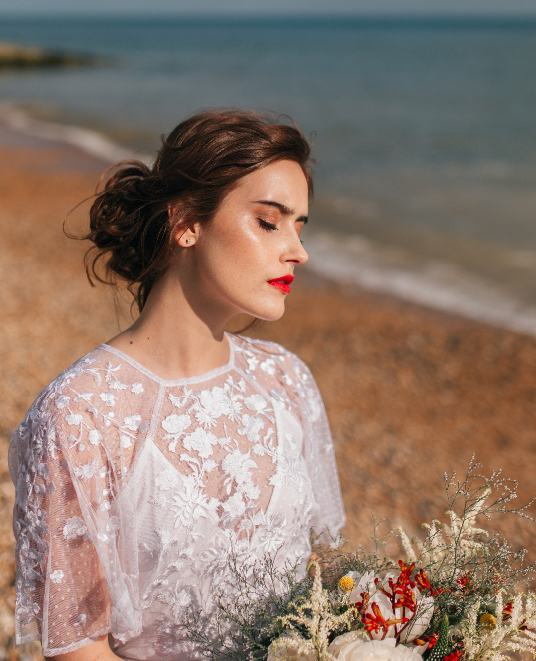 A woman with brown hair styled in an updo, wearing a white embroidered lace dress, standing on a beach holding a bouquet of flowers, with her eyes closed.