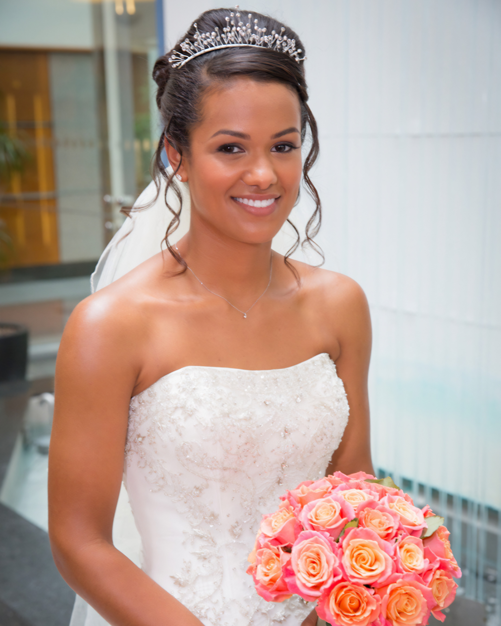 A smiling bride wearing a strapless wedding dress with floral embroidery, a tiara, and holding a bouquet of pink and peach roses.