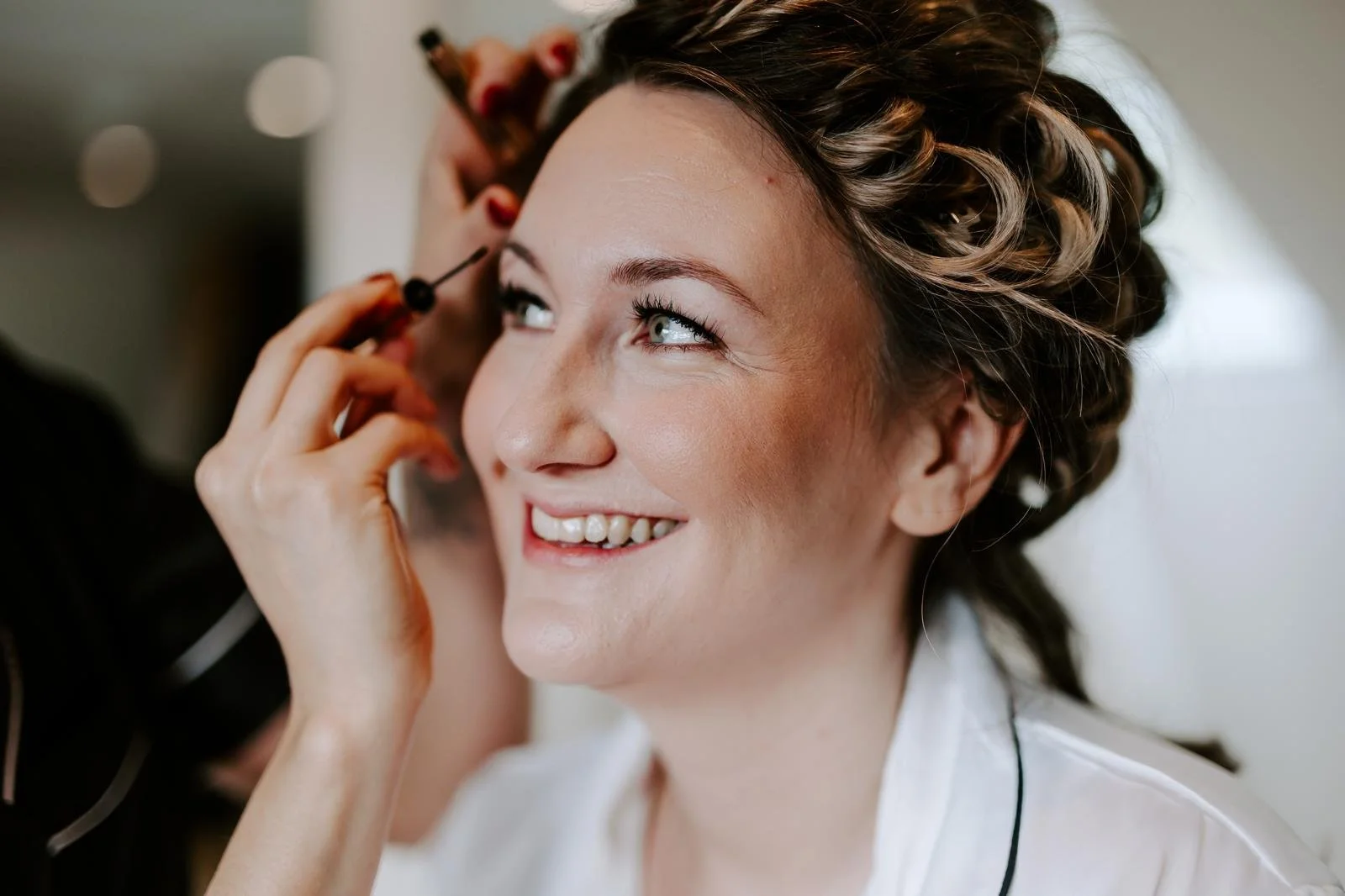 A woman with short wavy hair smiling as a makeup artist applies eye makeup with a brush.
