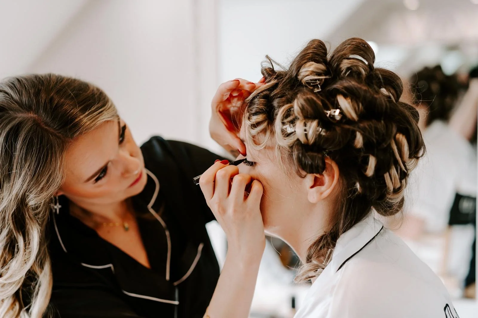 A woman with curled hair and hair rollers getting her makeup done by a makeup artist in a salon.