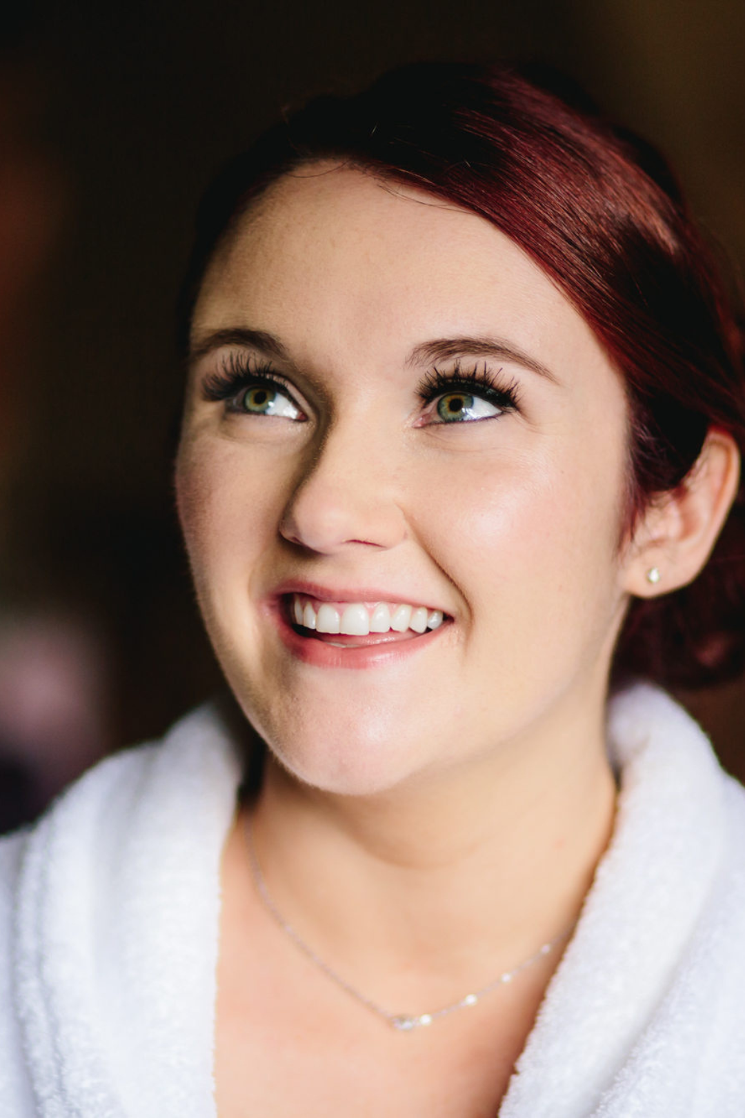 Close-up of a smiling young woman with red hair and green eyes, wearing a white bathrobe and small earrings.