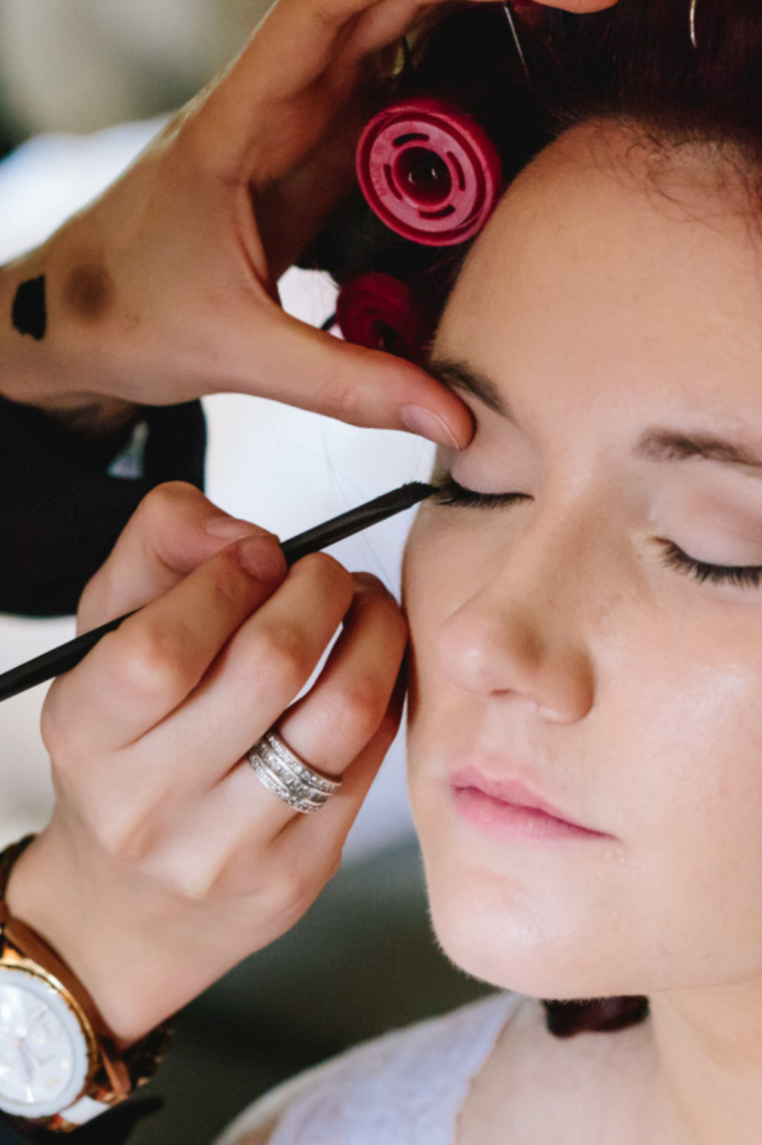A makeup artist is applying eye makeup to a woman with closed eyes, using a brush. The woman has long dark hair, and the artist has a tattoo on their finger and is wearing a watch and rings. The artist is also adjusting a pink hair roller or curlers in the woman's hair.