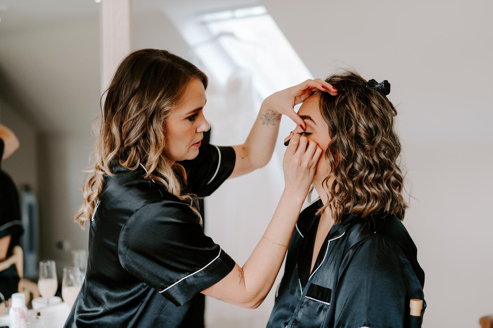A makeup artist applies makeup to a woman with curly hair in a room with white walls and natural light from skylights.