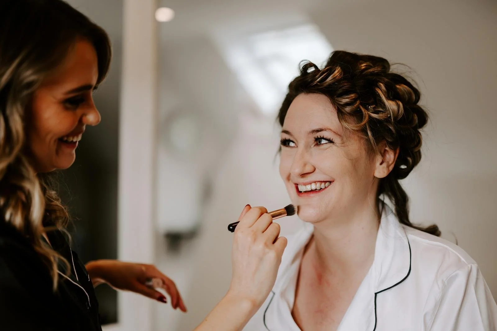 A woman with curled hair is getting her makeup done by another woman. The woman with curled hair is smiling and looking at the makeup artist, while the makeup artist is smiling and holding a makeup brush near her face.