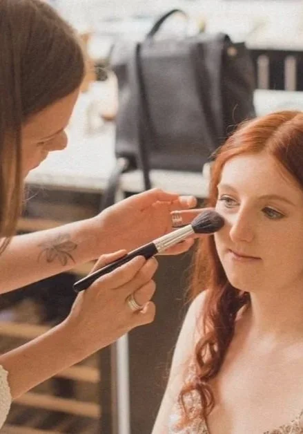 A makeup artist applying makeup to a young woman's face with a brush in a studio or backstage setting.