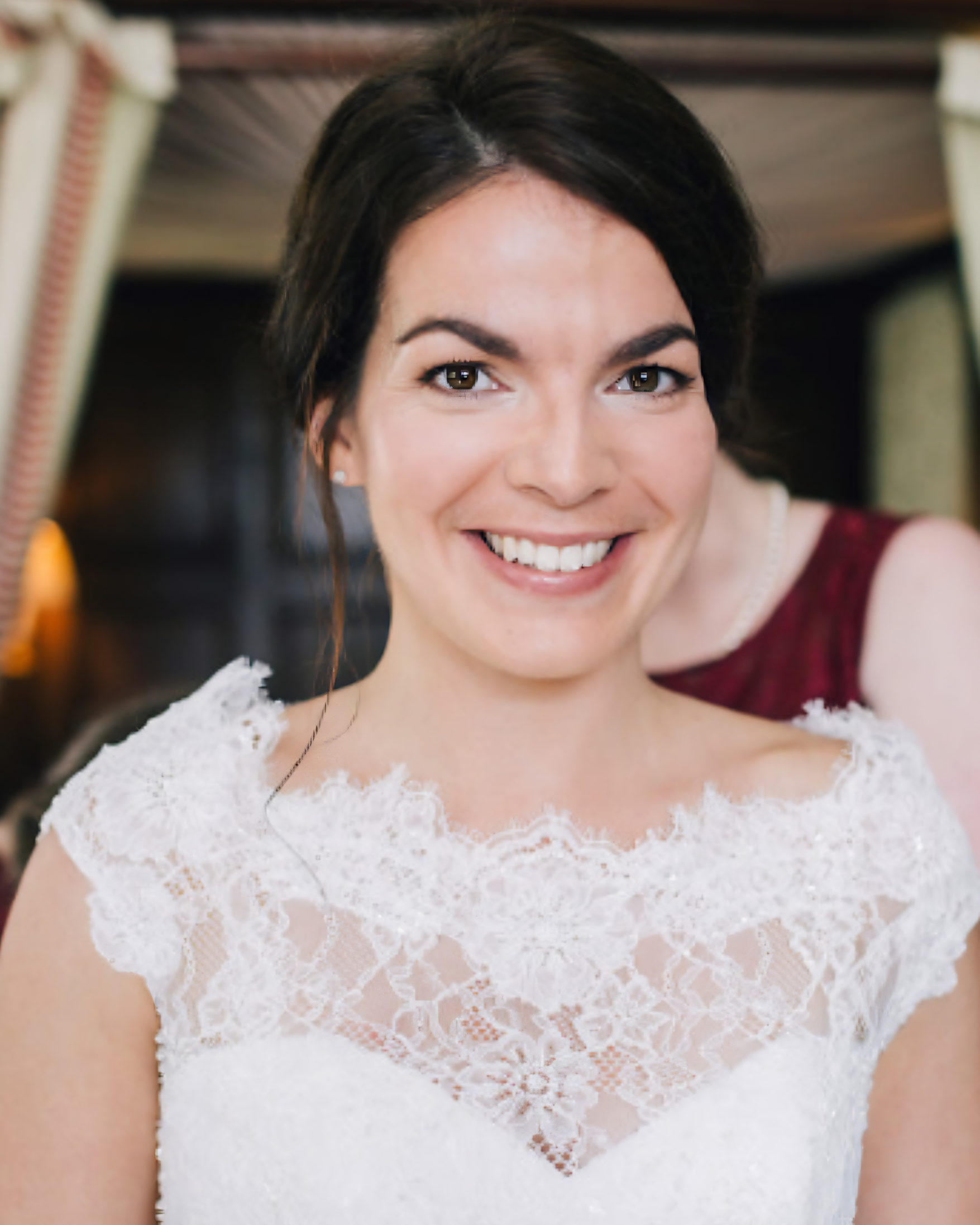 A woman with dark hair and a bright smile, wearing a white lace dress, indoors with blurred background.