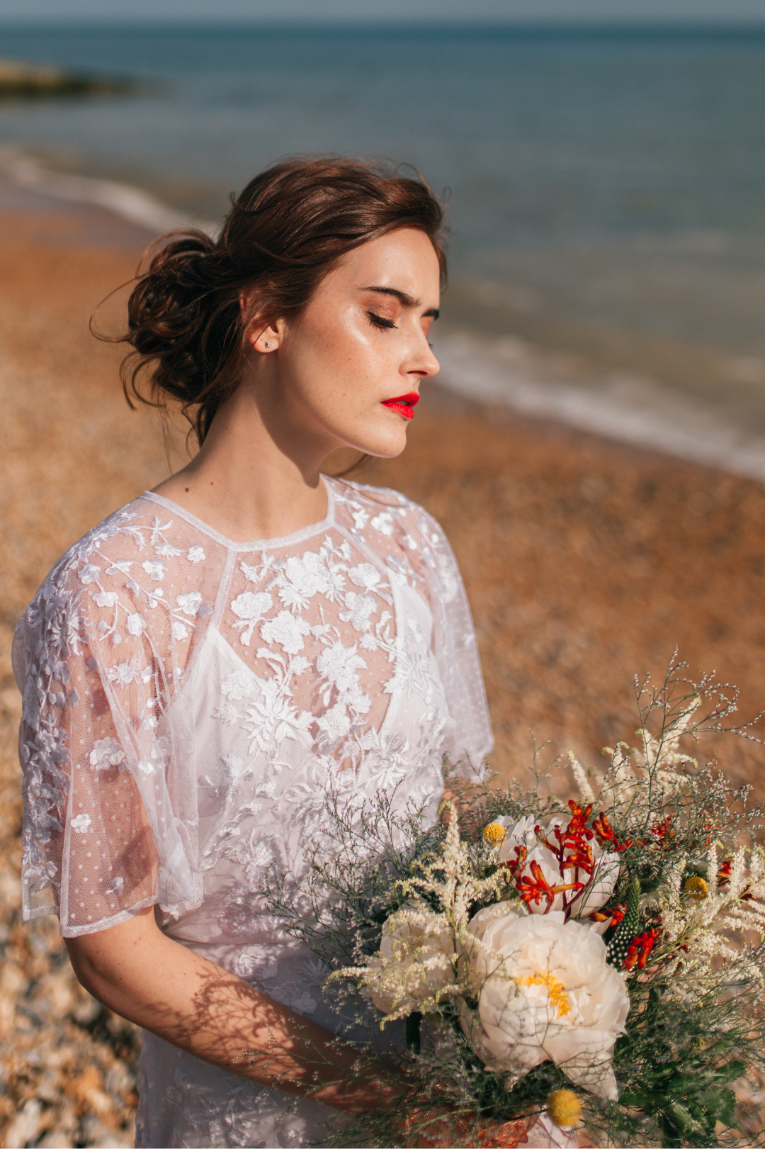 A woman in a white embroidered dress holding a bouquet of flowers on a beach with water in the background.
