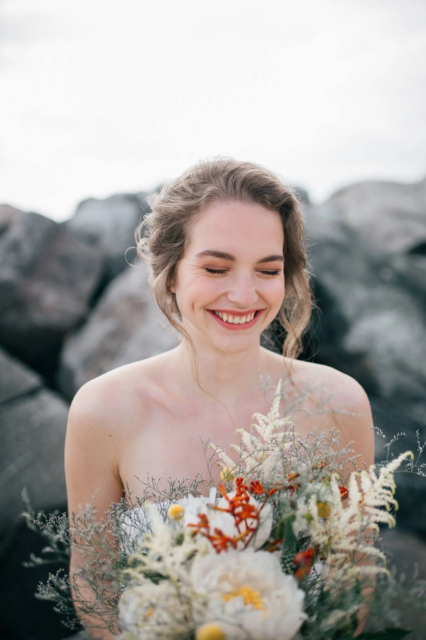 A young woman with curly hair and a bright smile holding a bouquet of various colorful flowers, standing outdoors with rocky background and an overcast sky.