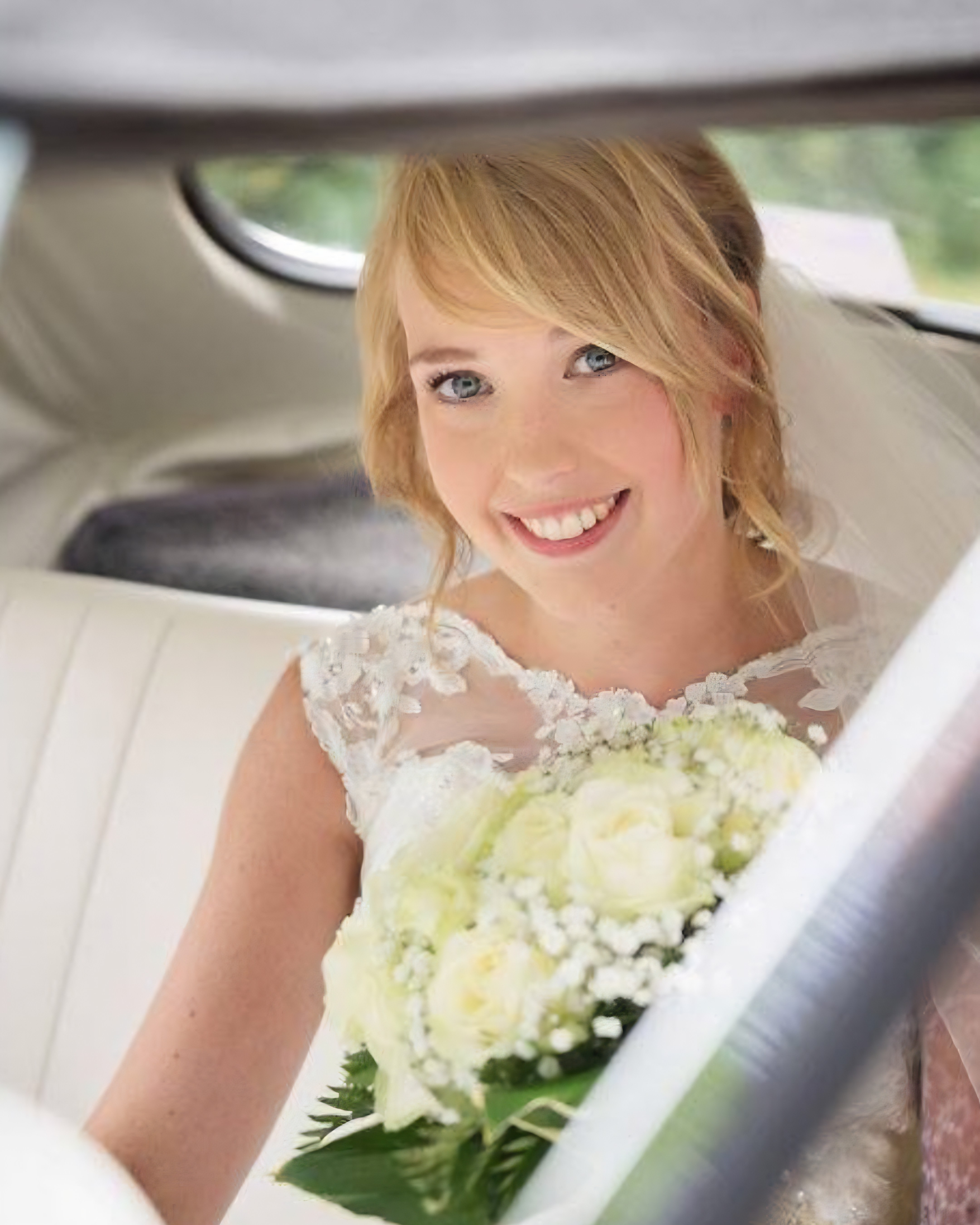 A woman with blonde hair and blue eyes smiling, dressed in a white lace dress, holding a bouquet of white flowers, sitting in a vehicle with a blurred background.