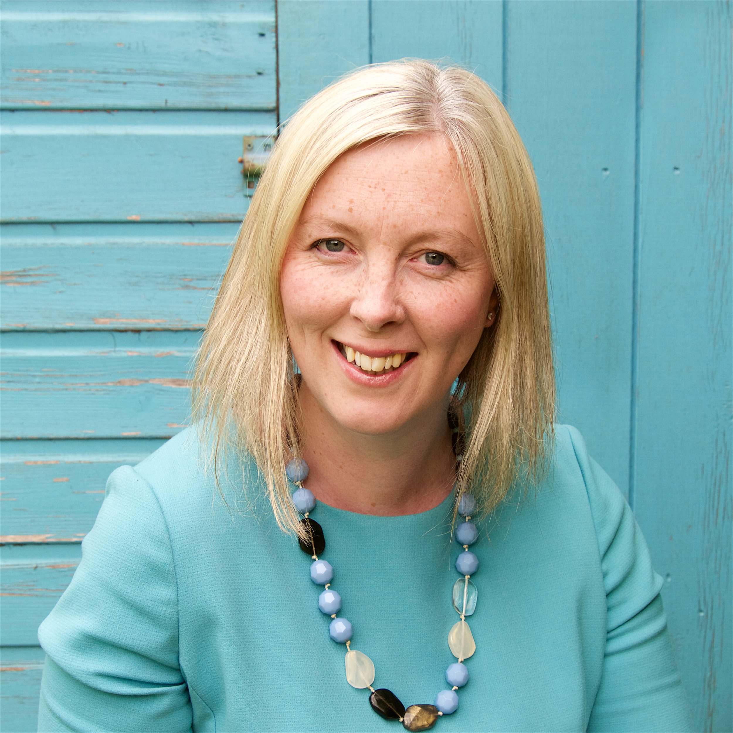 A woman with blonde hair wearing a turquoise blouse and a beaded necklace, smiling in front of a blue wooden background.