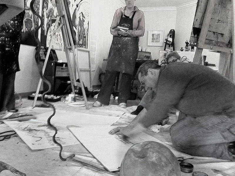 People working in an art studio with paintings and artworks on the walls and easels, one person is kneeling on the floor working on a large piece of paper, others are standing observing.