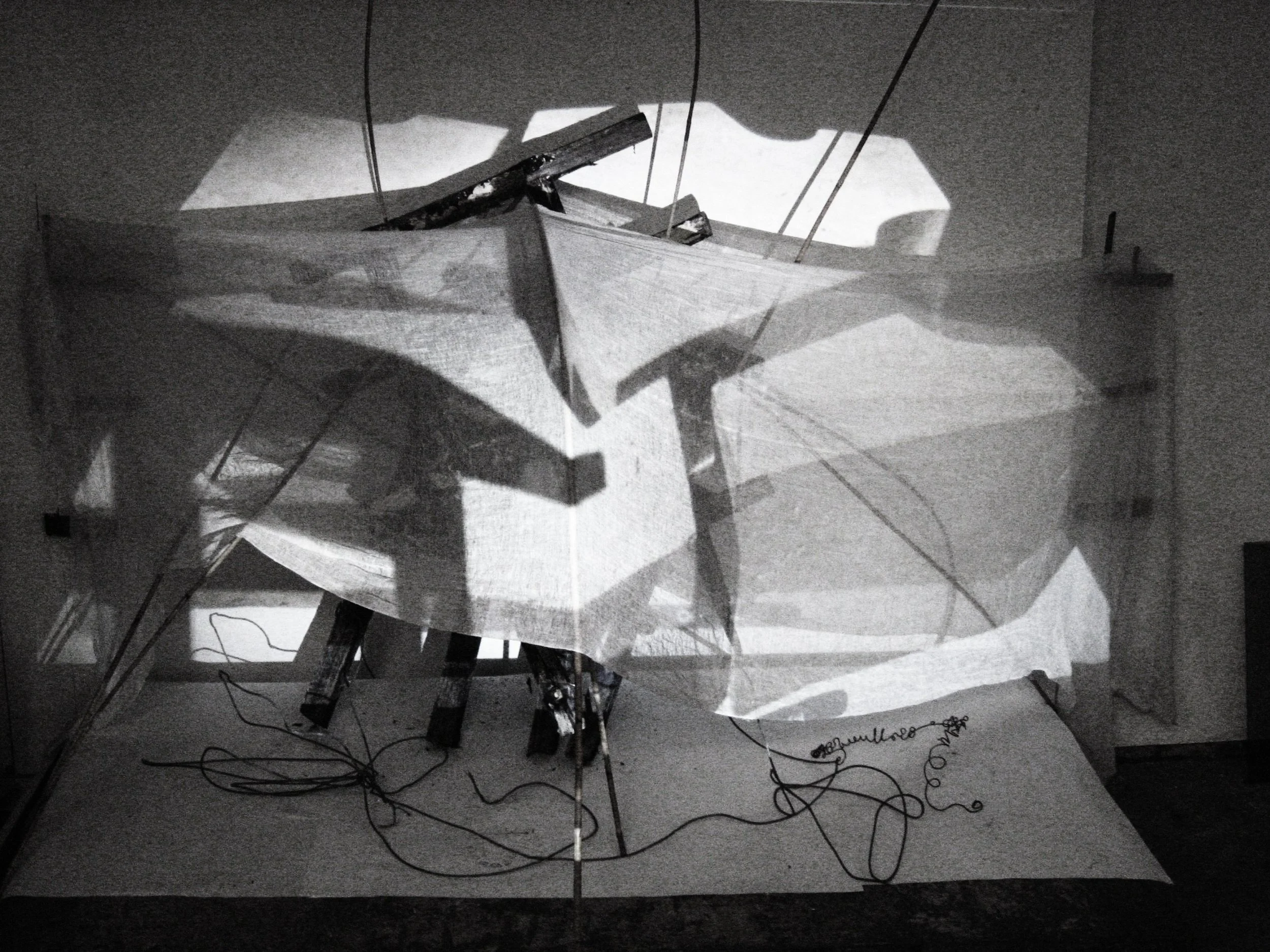 An art installation featuring a large, irregularly shaped sheet or fabric suspended in the air, illuminated with shadows, with wooden and wire components attached to a platform on the ground.
