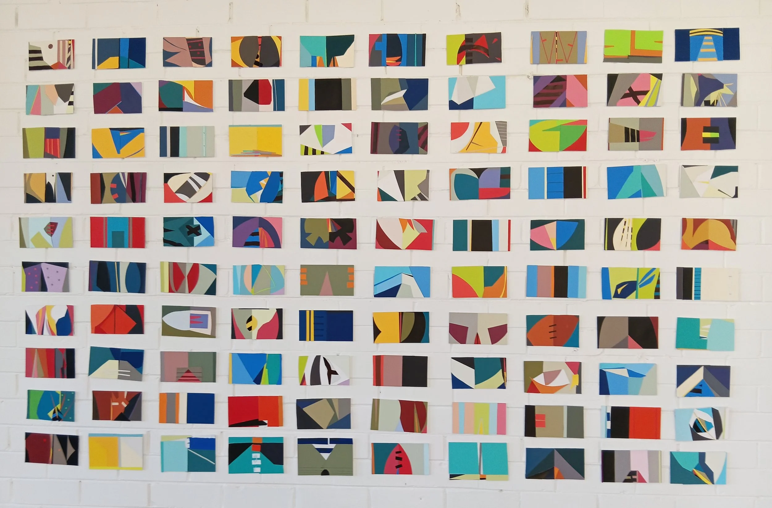 An art display featuring numerous small abstract paintings arranged in rows on a white brick wall. The paintings are colorful and geometric, with various shapes and lines.