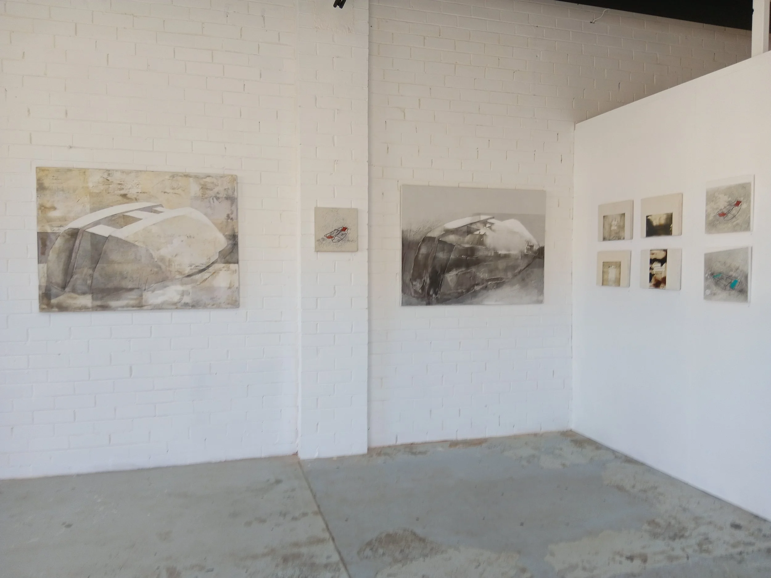 Art gallery with abstract paintings and small mixed media artworks on white brick walls.