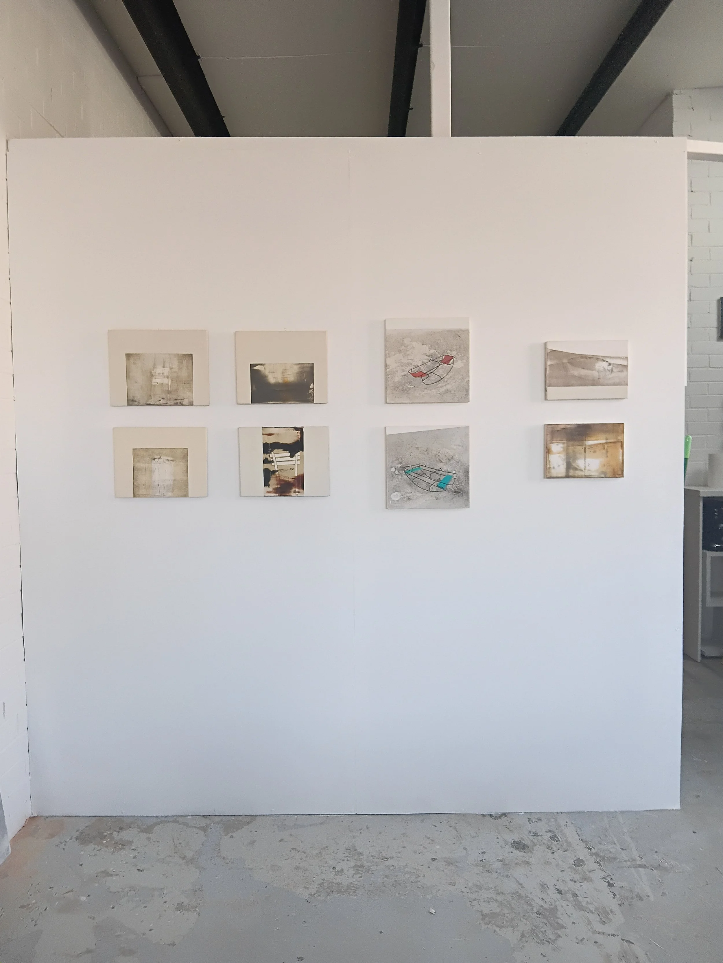 Art gallery display with eight small abstract paintings on a white wall, in an industrial space.