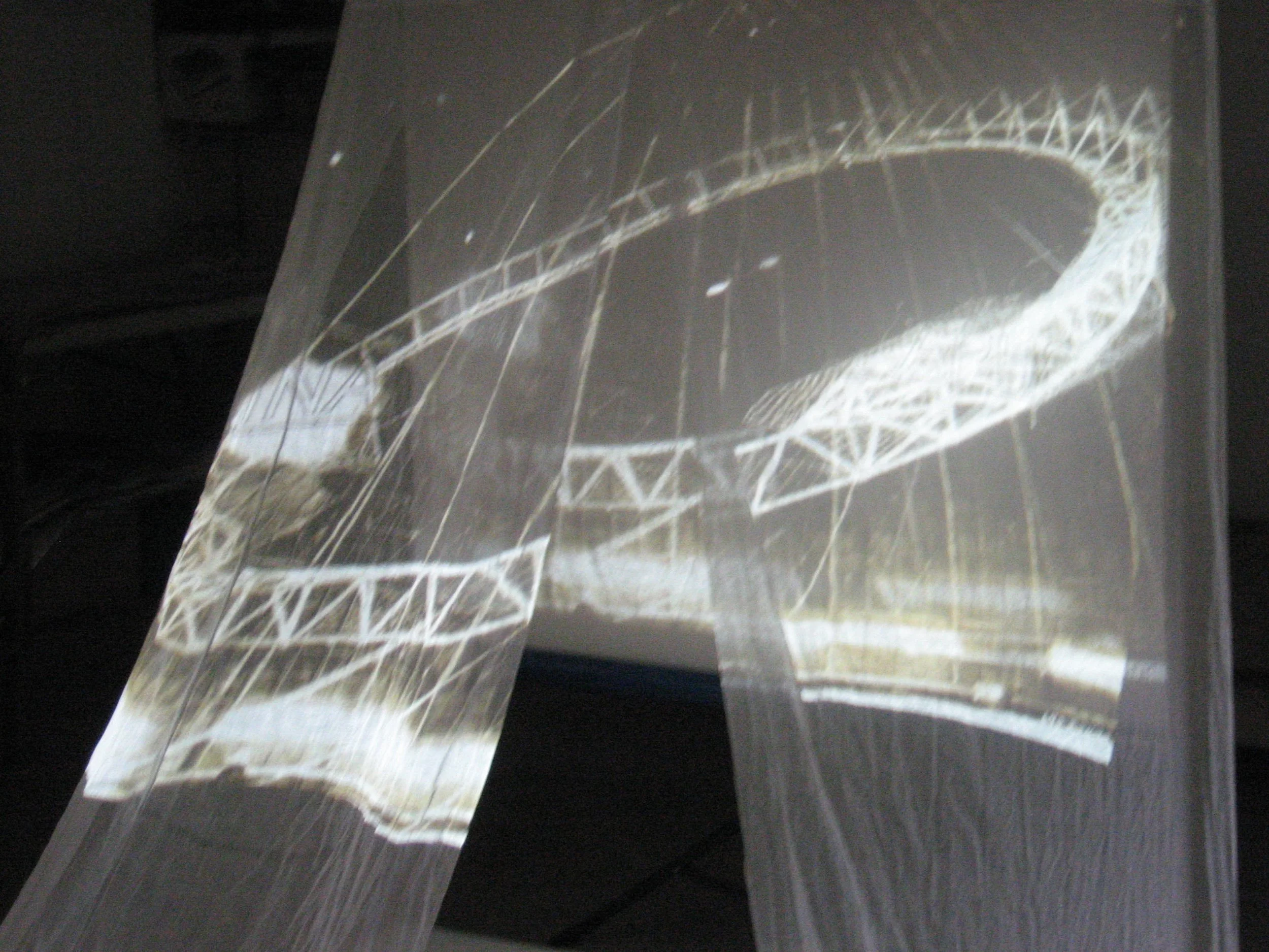 Photograph of a large white suspended structural framework with a central circular opening, seen through a semi-transparent sheet with reflections.