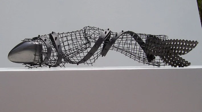 Abstract sculpture resembling a fish made of metal and wire, with a streamlined head and a tail with round patterns, displayed on a white platform.