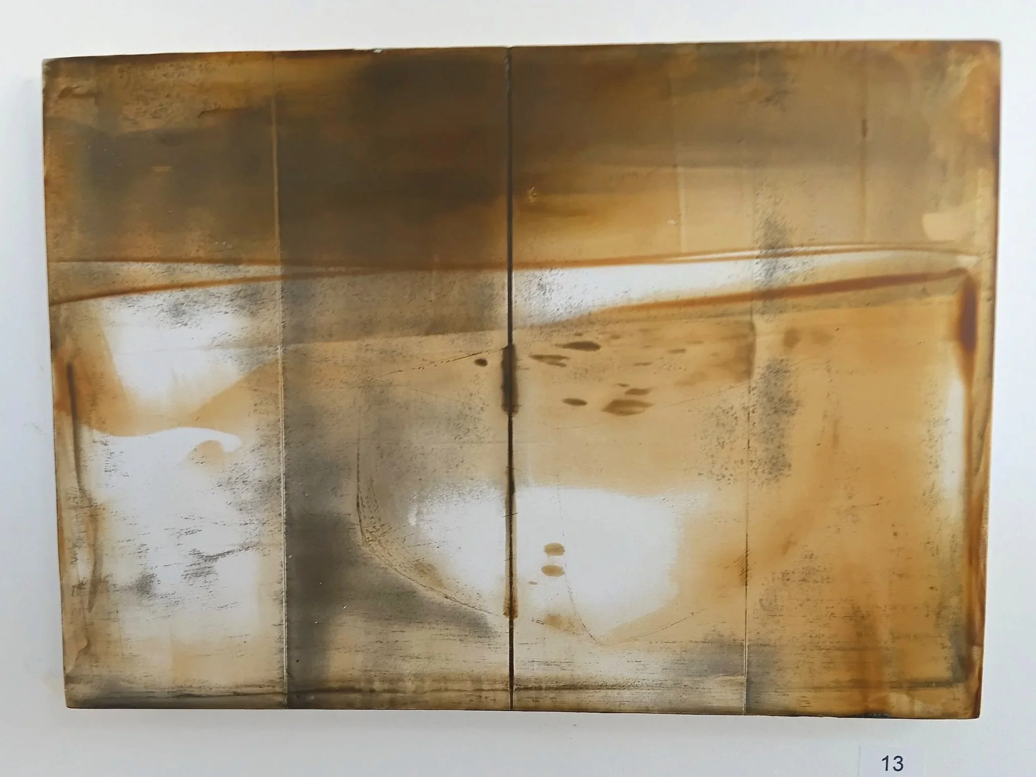Abstract mixed media artwork featuring metallic, rust, and beige tones with irregular patterns and reflections, mounted on a white wall with a small label marked '13'.