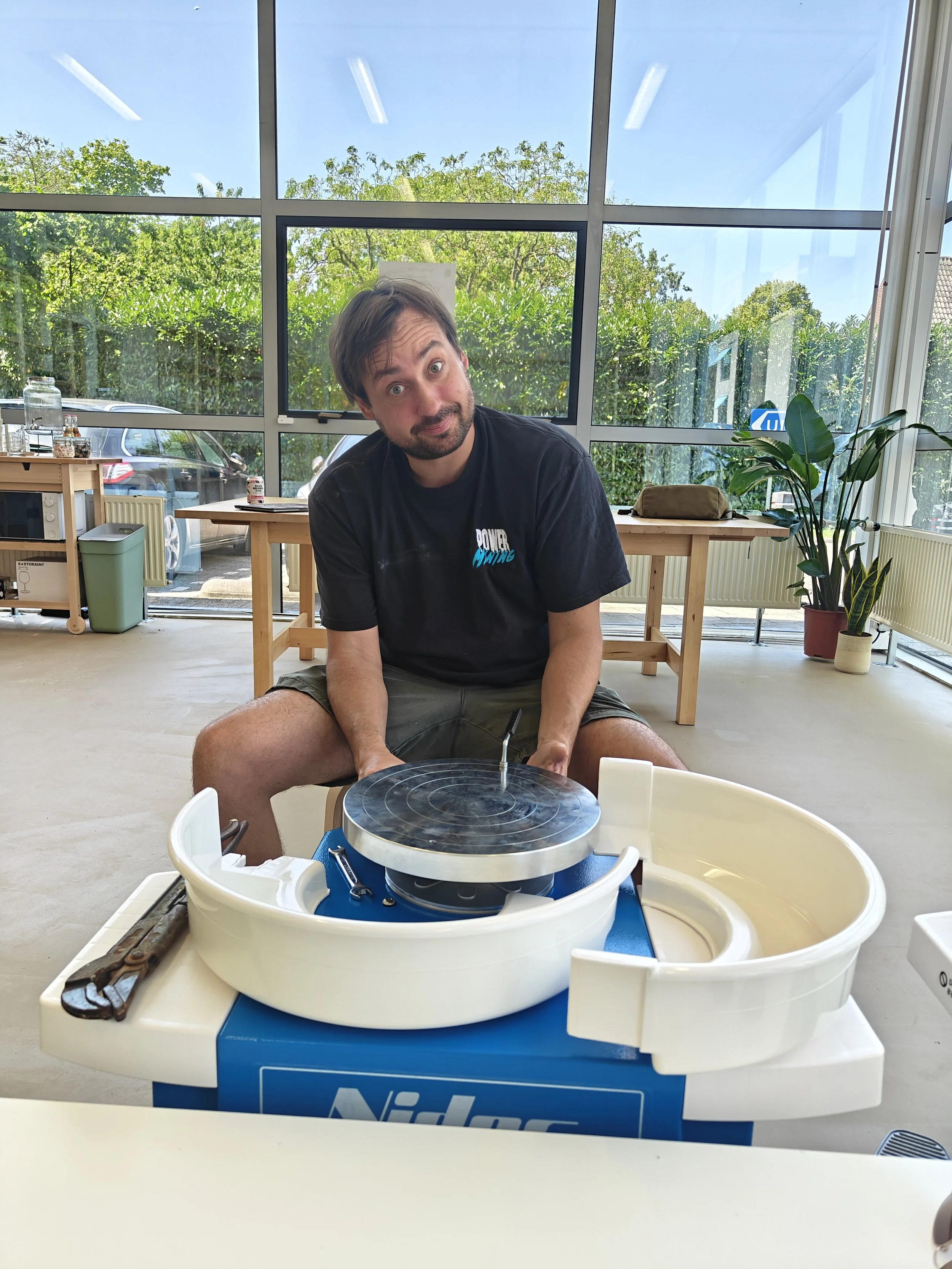 Man met een lach zit achter een keramiek draaibord in een lichte kamer met grote ramen en groene planten.