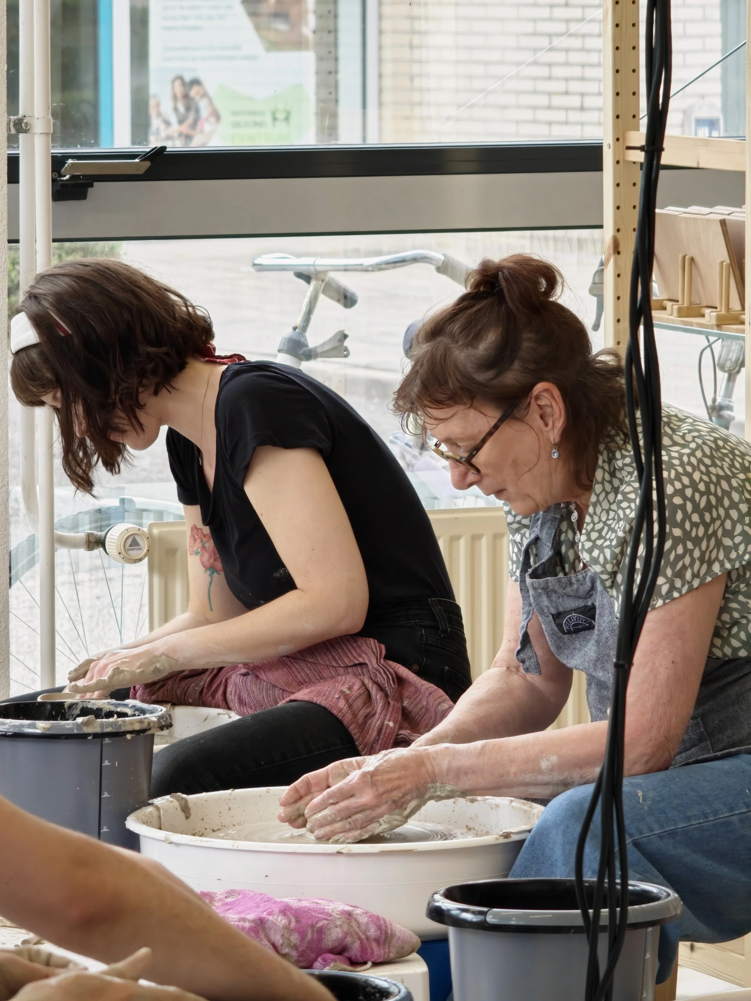 Twee vrouwen die handweven met vaten en lichte kleuren in een ruimte met grote ramen.