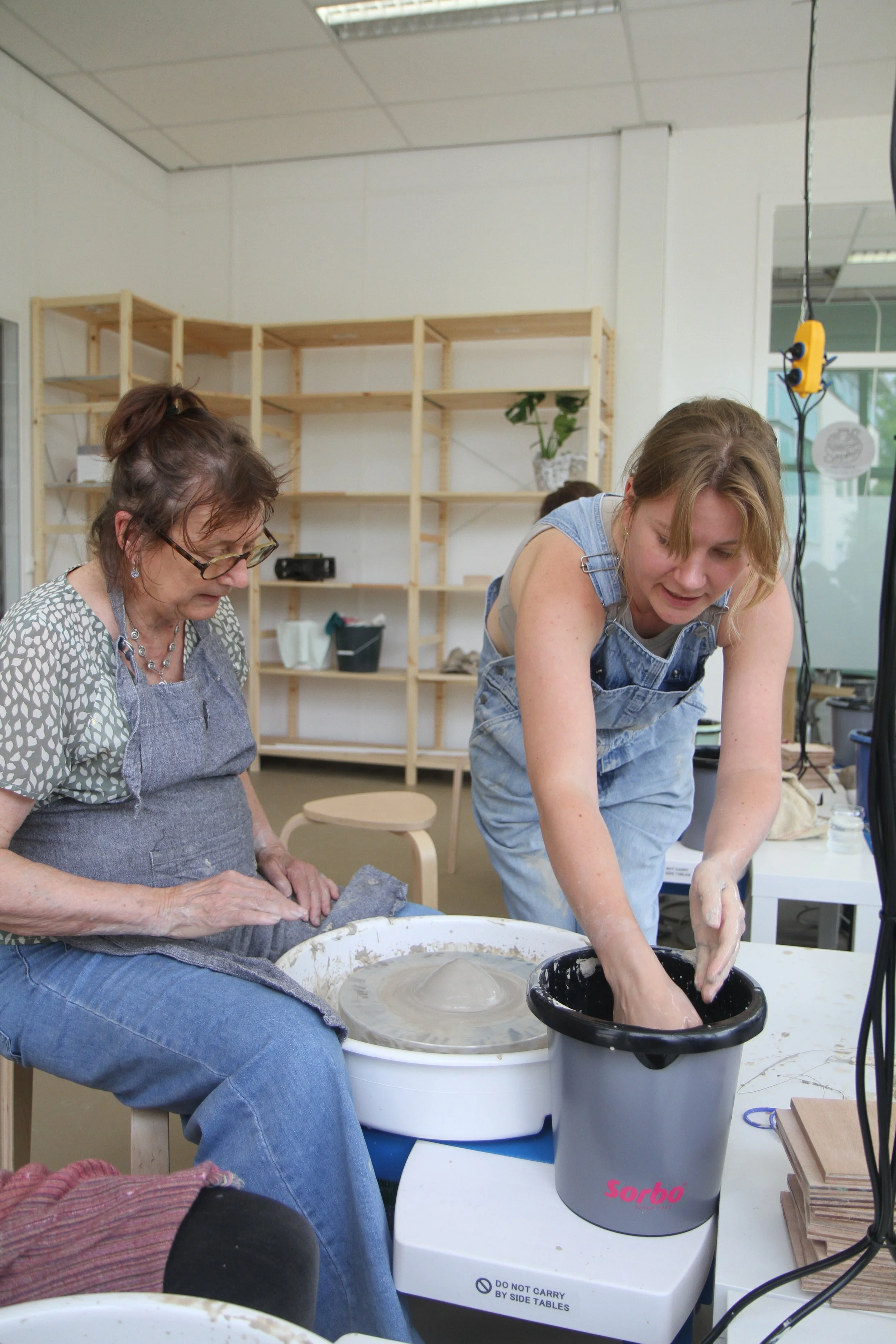 Twee vrouwen werken samen aan keramiek op een tekentafel, een van hen rolt klei uit op een draaischijf, de ander kijkt toe.