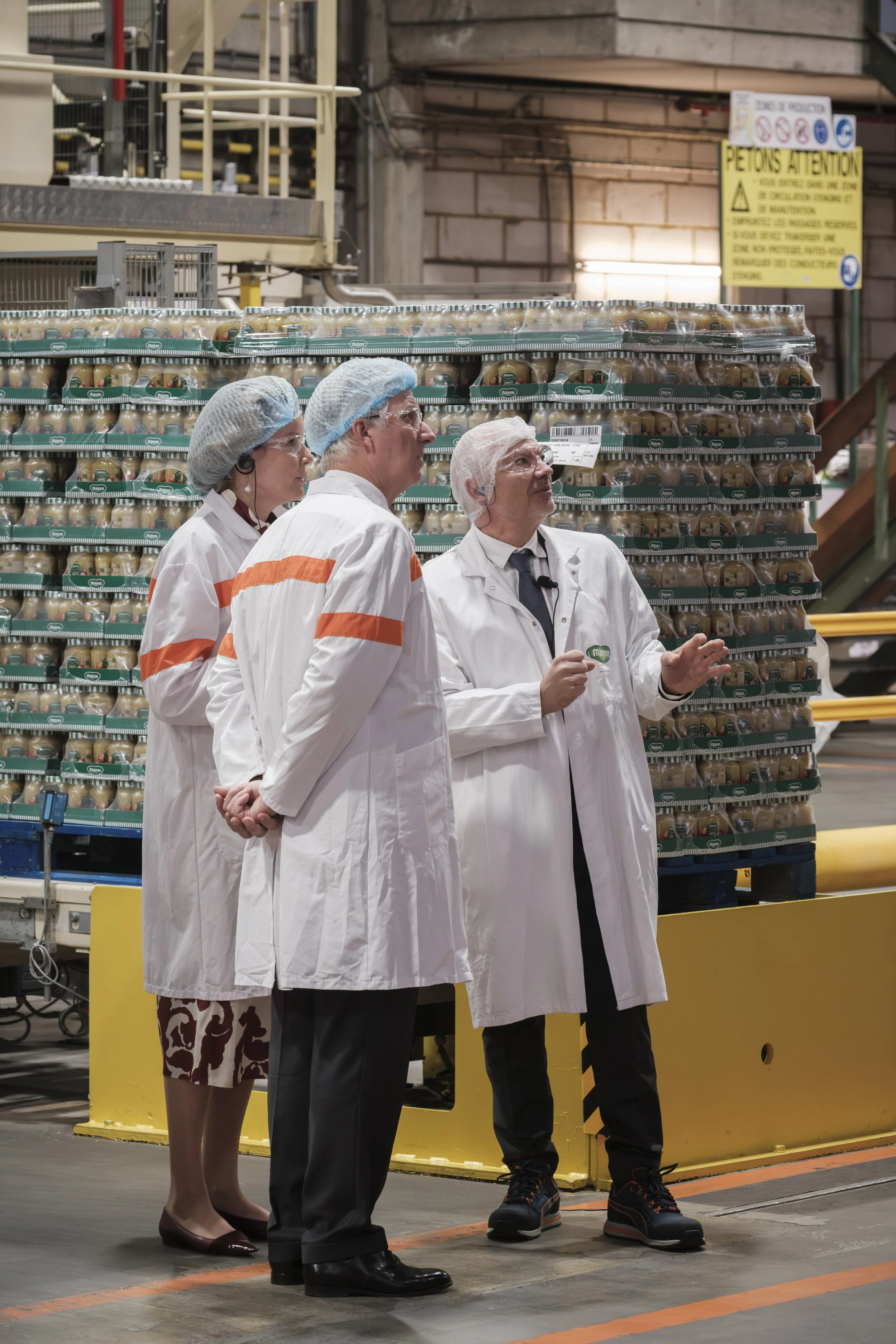 Le roi Philippe et la reine Mathilde en tenue de protection dans l'usine Materne, présente sur fond de palettes de produits emballés. | QSphotography | Namur