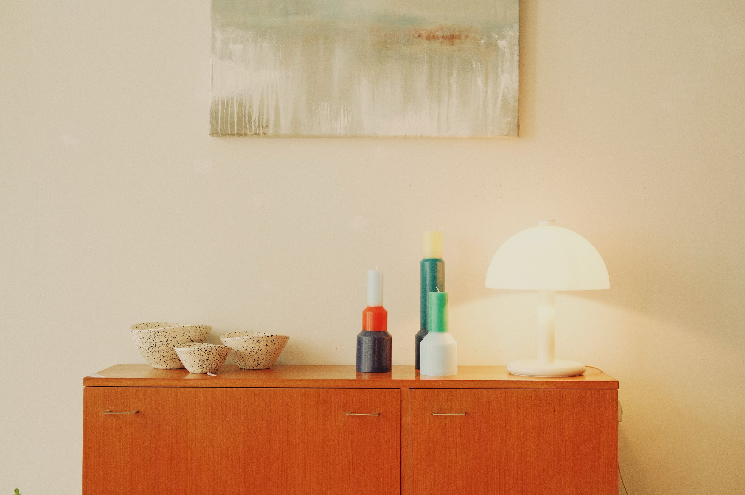 Une commode en bois avec plusieurs bols en céramique à gauche, trois bougies colorées avec supports de différentes hauteurs au centre, et une lampe de bureau blanche à droite, contre un mur beige. | QSphotography | Namur