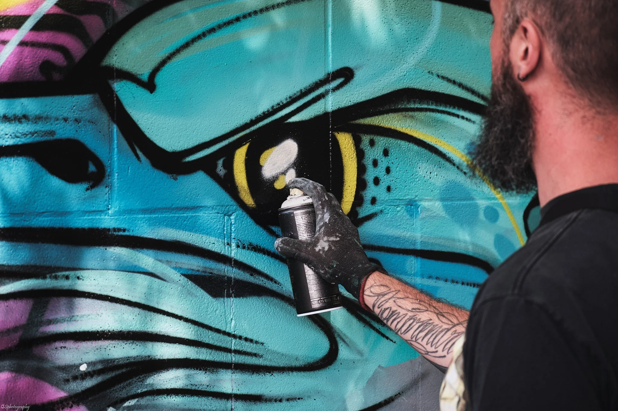 Un homme avec une barbe et un tatouage sur le bras, portant un gant noir, utilise une bombe de peinture en spray pour créer un graffiti d'un visage de style bande dessinée sur un mur coloré.