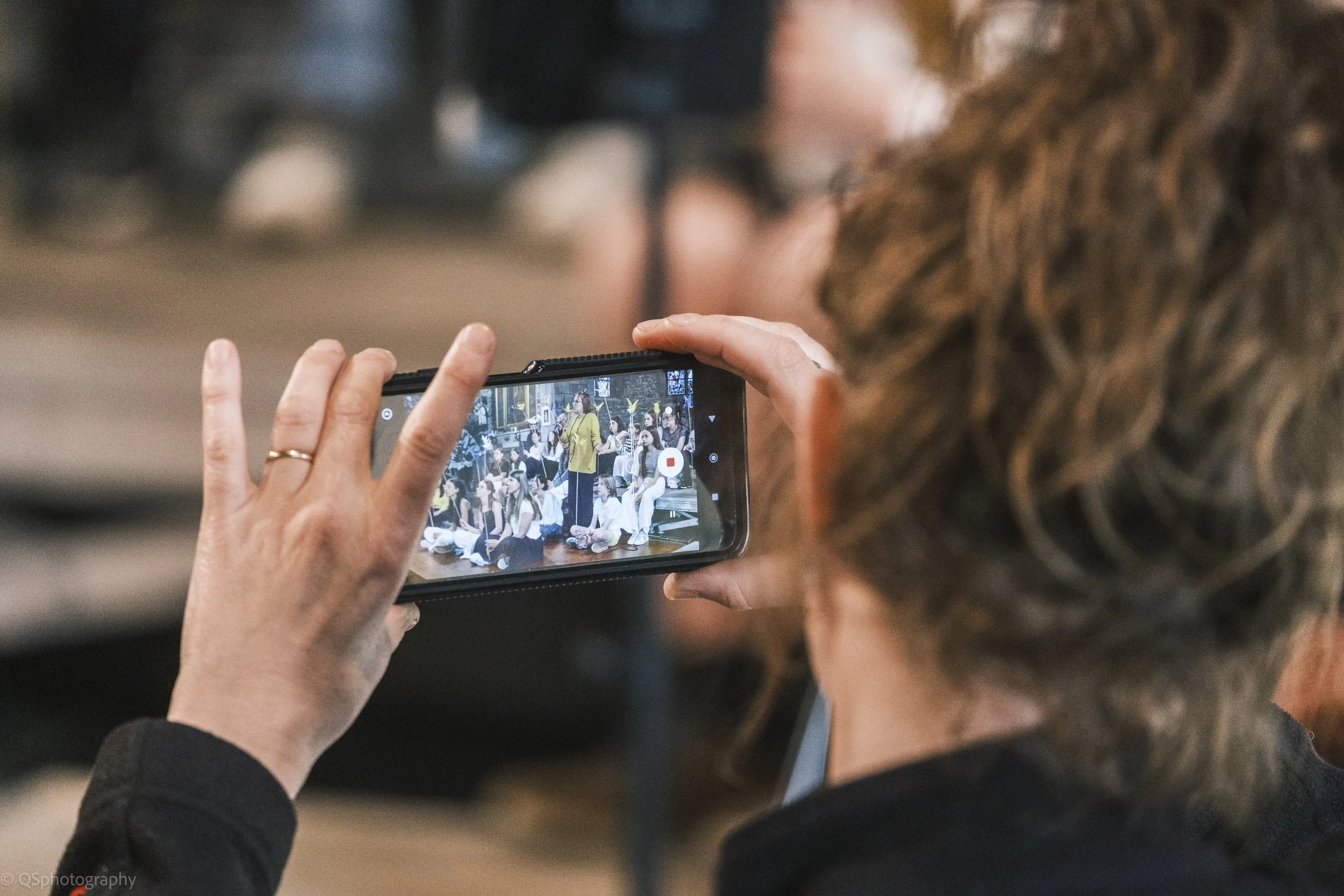 Une personne tient un téléphone portable, enregistrant ou prenant une photo d'un groupe de personnes sur scène à l'intérieur.