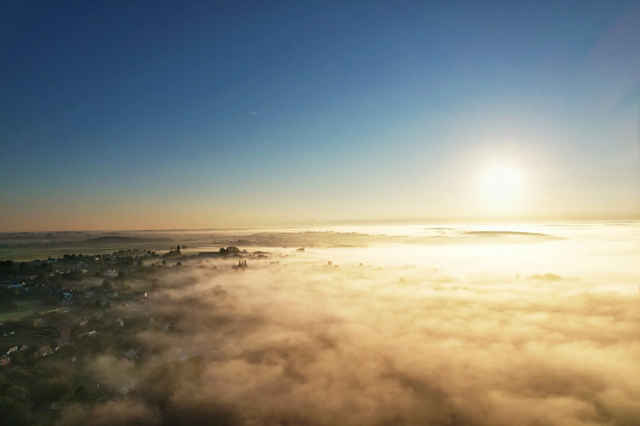 Paysage avec des maisons et des arbres couverts de brouillard au lever du soleil, ciel clair avec un petit avion passant. | QSphotography | Namur