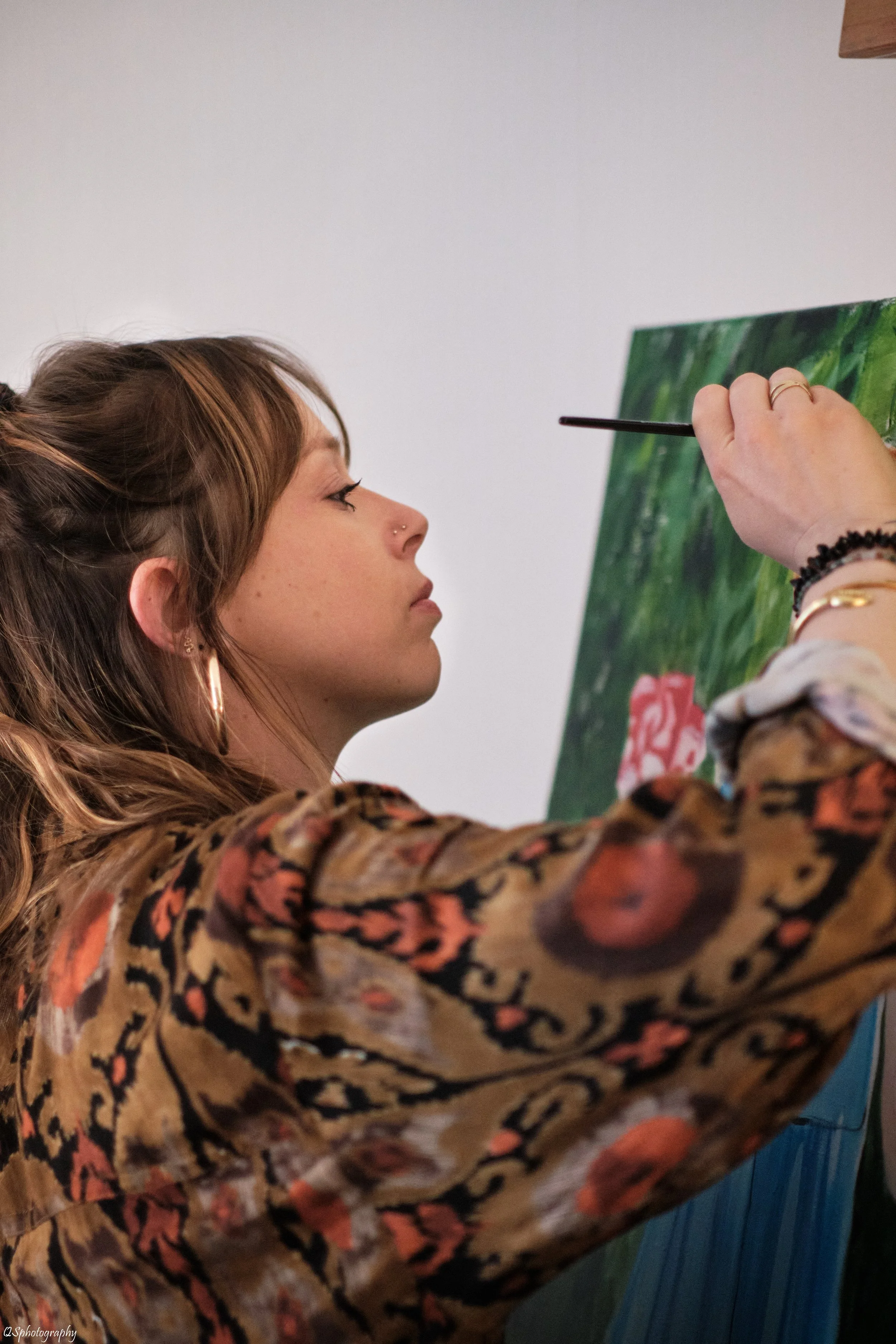 Une femme avec des boucles d'oreilles en or peignant une toile avec un pinceau. Elle porte une robe imprimée avec des motifs floraux ou animaux. | QSphotography | Namur