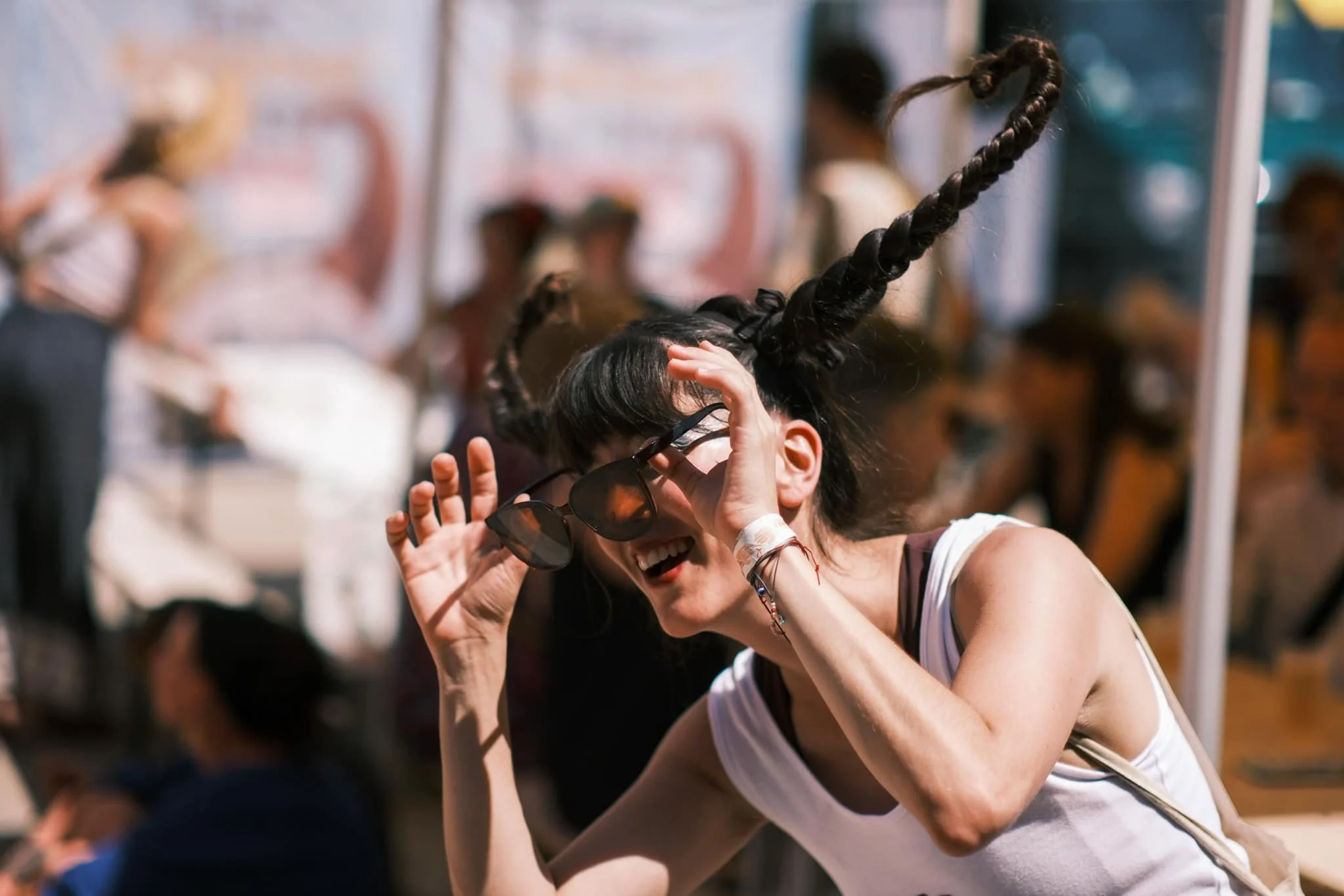 Une jeune femme avec des cheveux tressés en forme de cornes, portant des lunettes de soleil, sourit lors d'un événement social en plein air. | QSphotography | Namur