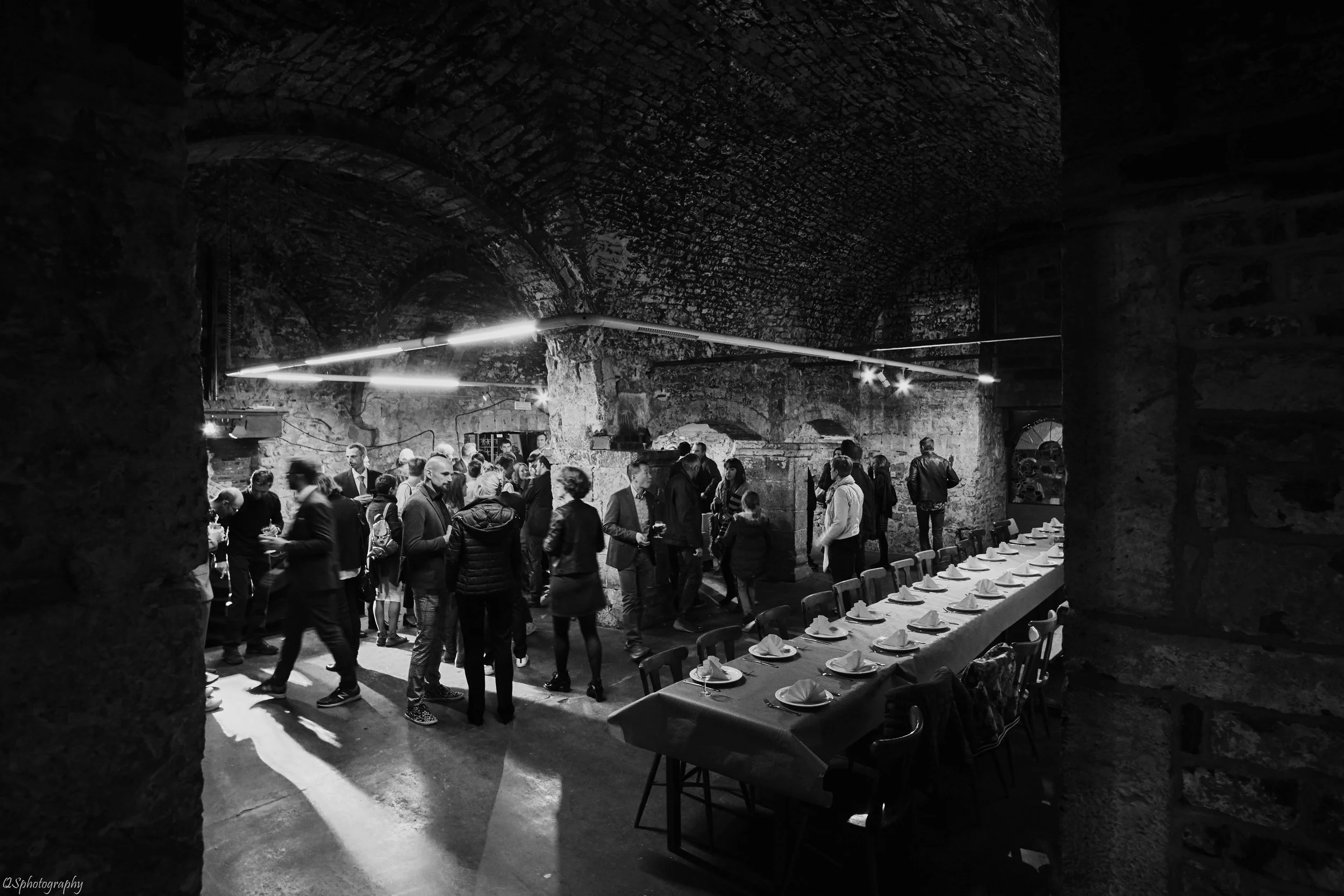 Une image en noir et blanc d'une réception organisée dans un espace industriel avec des murs en pierre et un plafond en arc, des invités en costume discutant, une longue table dressée avec des assiettes et des serviettes.| QSphotography | Namur