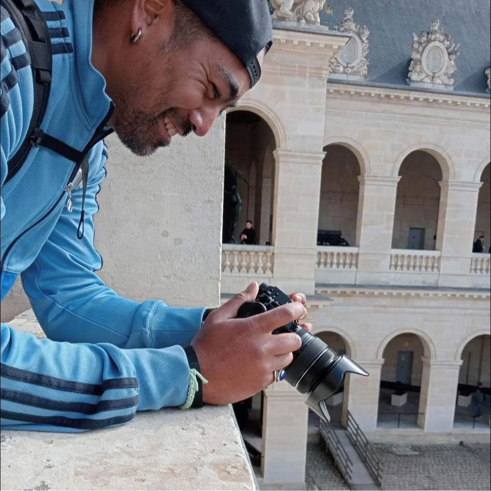 QSphotography souriant portant une veste bleue et un bonnet noir, tenant un appareil photo, se tenant près d'un balcon avec une architecture en pierre, dans un bâtiment historique, probablement en voyage ou en photographie. | QSphotography | Namur