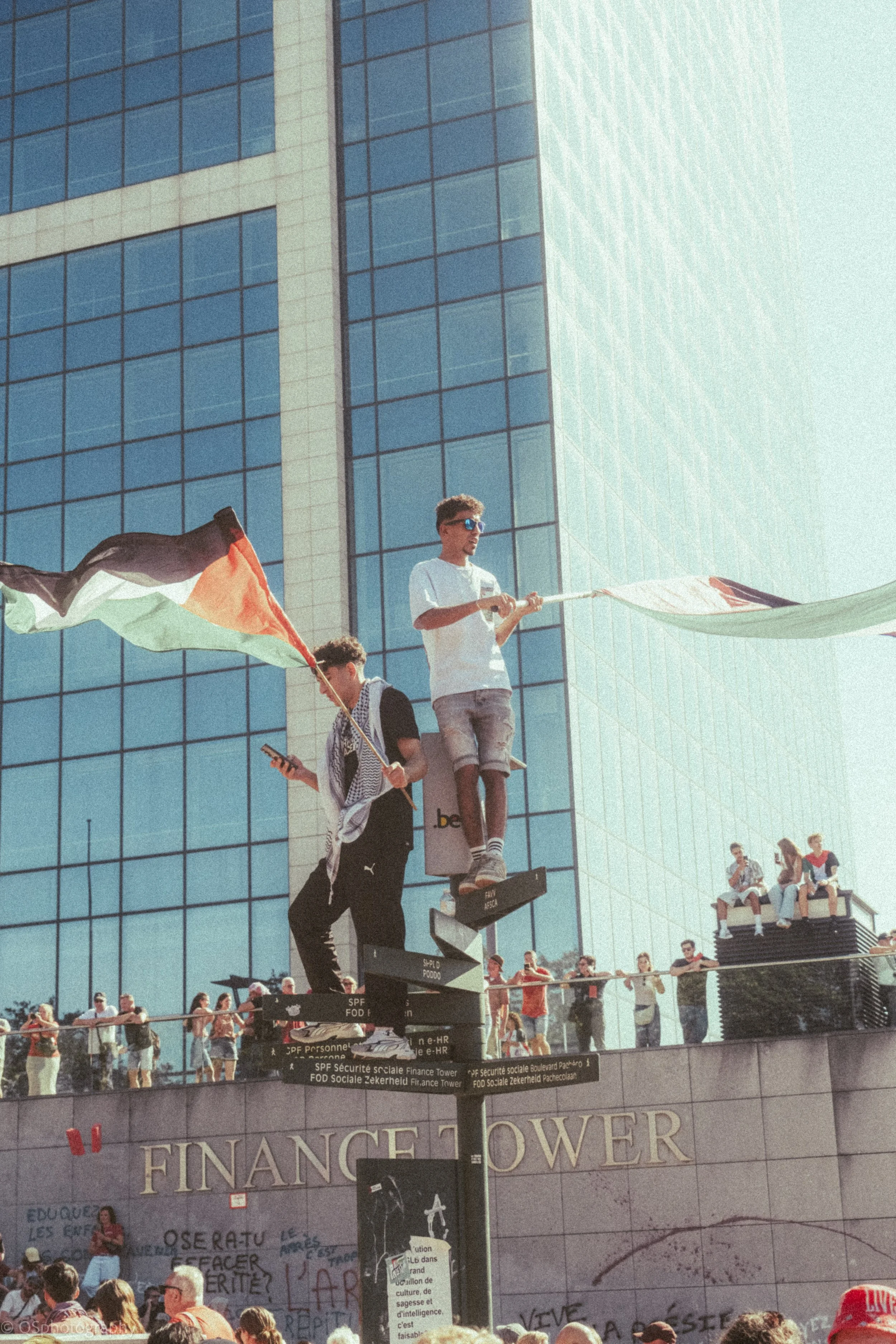 Deux hommes marchant sur un fil tendu au-dessus de la rue devant un grand immeuble en verre, brandissant un drapeau aux couleurs de la Palestine, avec beaucoup de personnes en arrière-plan, dont certaines assises sur un toit. | QSphotography | Namur
