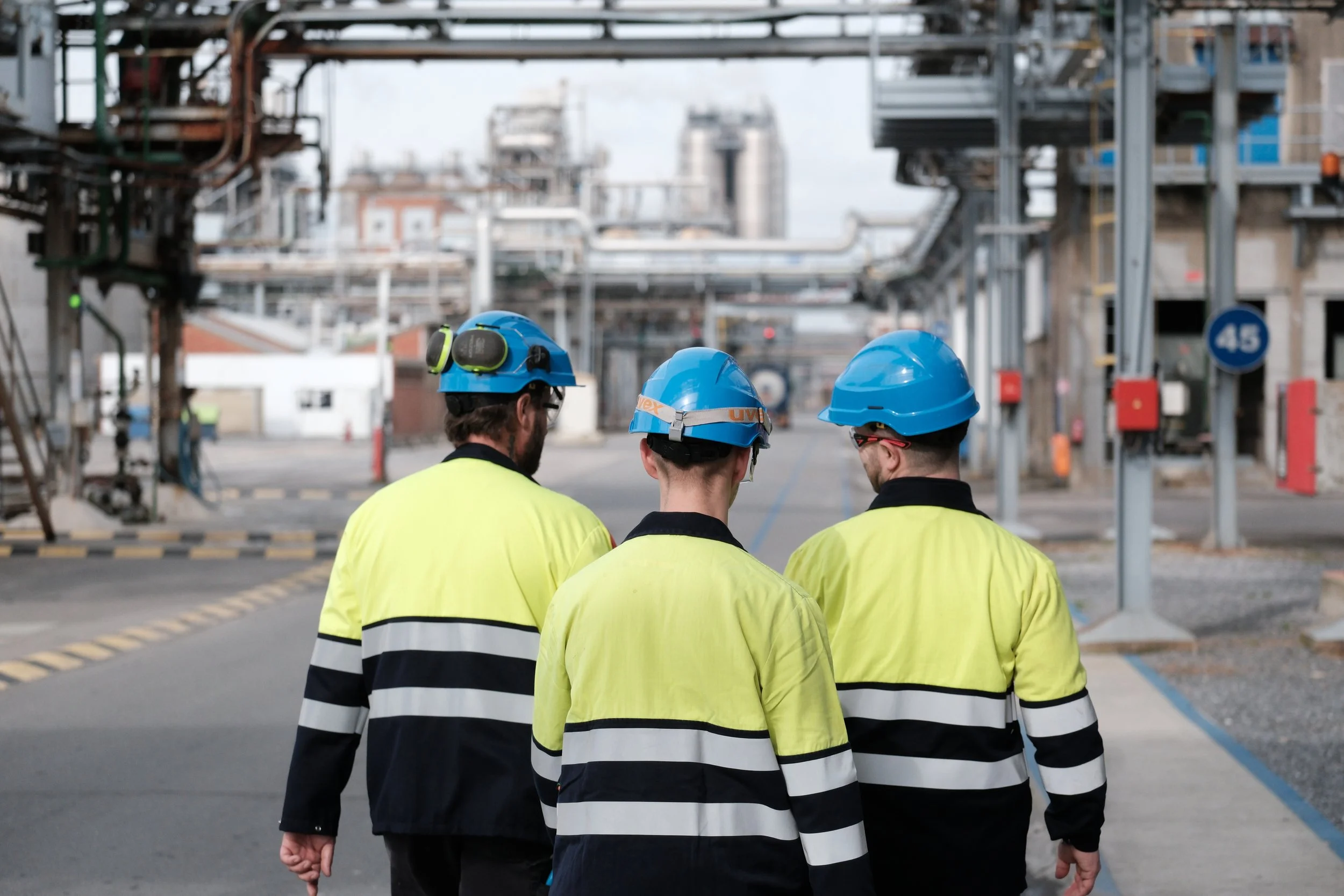 Trois travailleurs portant des casques bleus et des vestes de sécurité jaunes avec bandes réfléchissantes, marchant dans une zone industrielle, avec des structures métalliques et des bâtiments en arrière-plan. | QSphotography | Namur