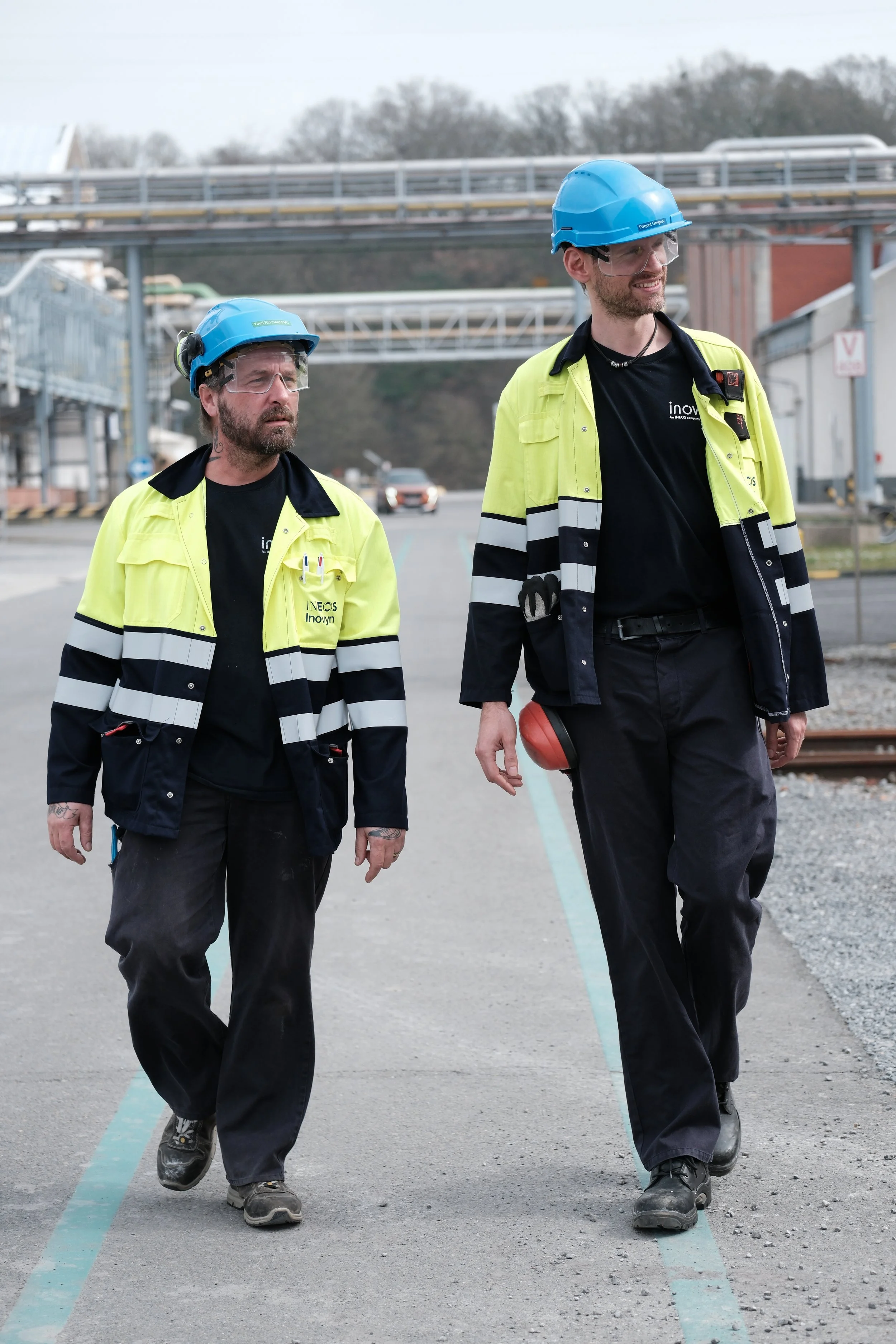 Deux ouvriers en vêtements de chantier avec casques de sécurité et gilets réfléchissants marchent sur un site industriel avec des voies ferrées de chaque côté. | QSphotography | Namur