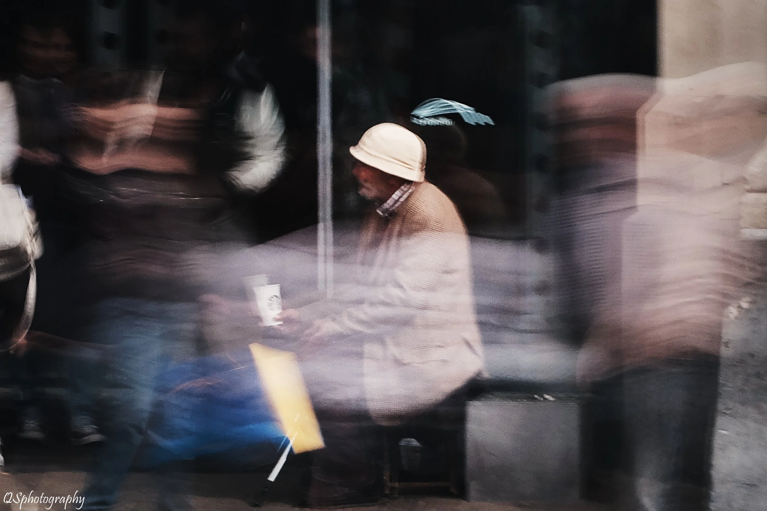 Un homme âgé avec un chapeau beige assis à une table dans une rue animée, flouté par le mouvement et la photo réflexe, entouré de passants. | QSphotography | Namur