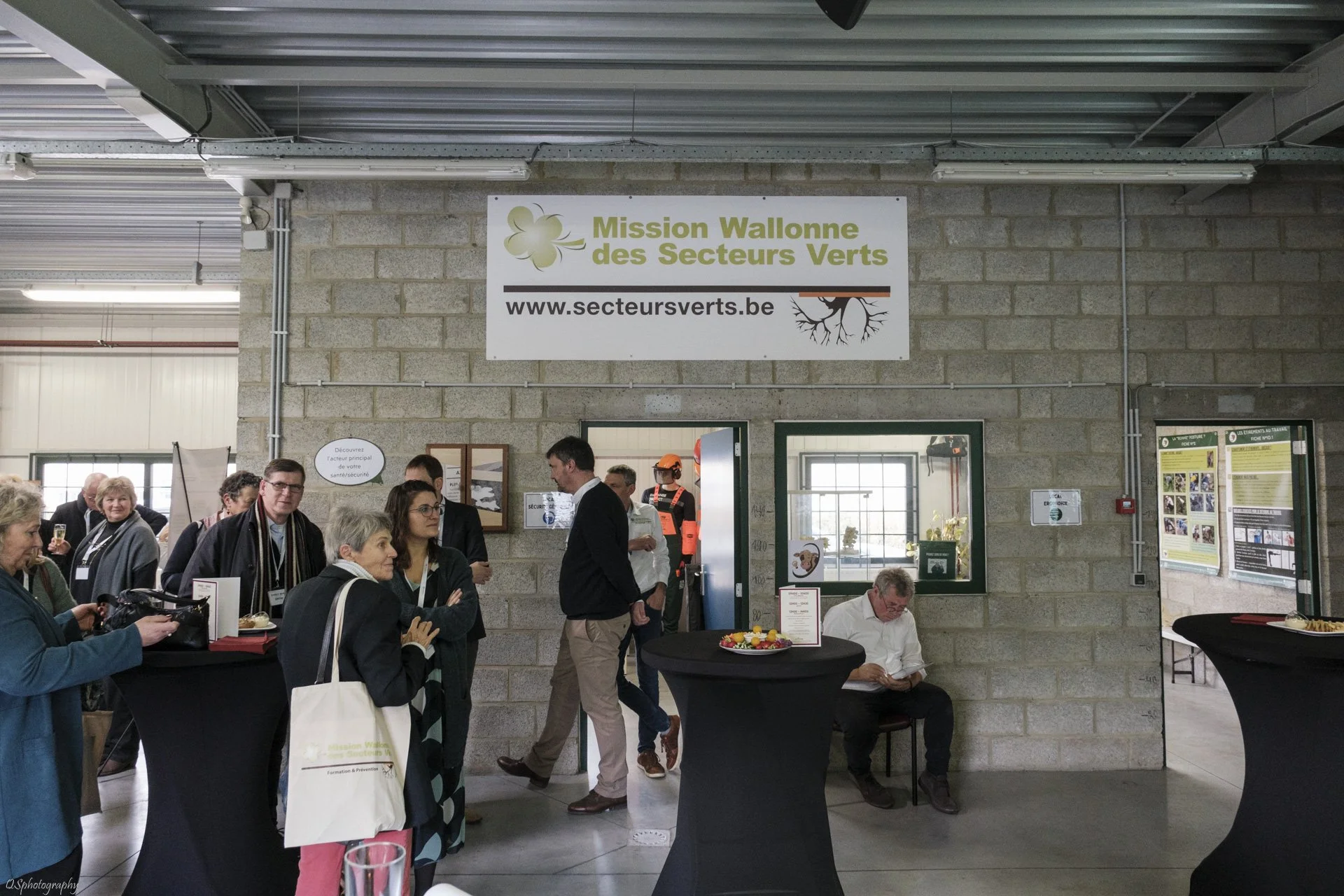 Un groupe de personnes rassemble autour de tables hautes lors d'un événement, avec un fond d'une paroi de briques, papier d'information, et un panneau annonçant la Mission Wallonne des Secteurs Verts. | QSphotography | Namur
