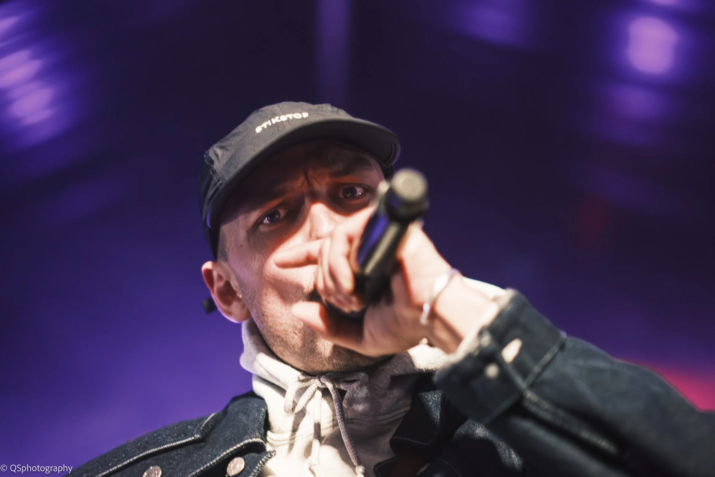 Un homme portant une casquette sombre et un sweat à capuche, tenant un microphone et regardant vers l'appareil photo, avec un fond bleu violet flou.