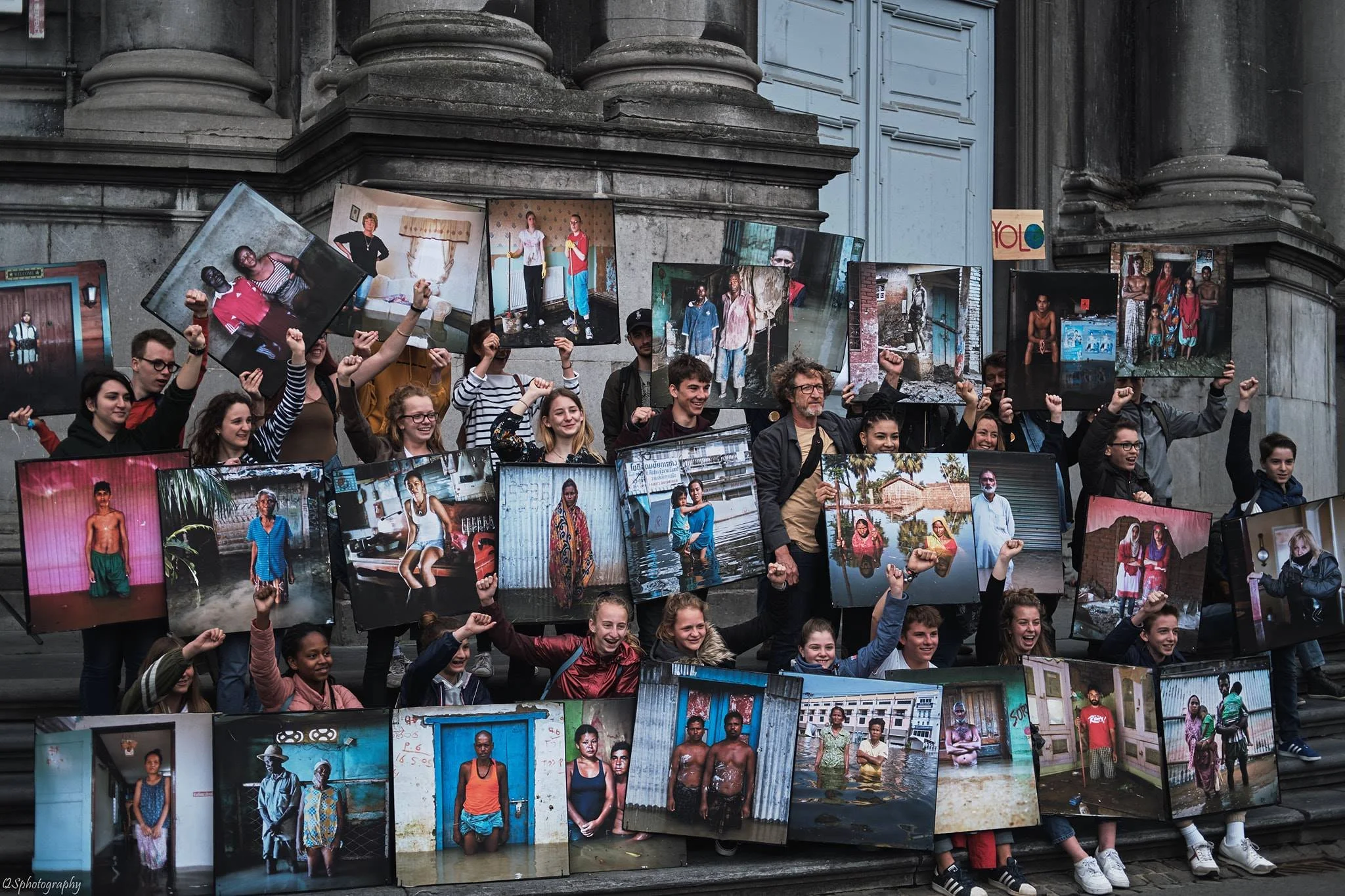 Groupe de personnes debout sur des escaliers tenant des panneaux avec des photos de différentes personnes dans divers environnements et situations.